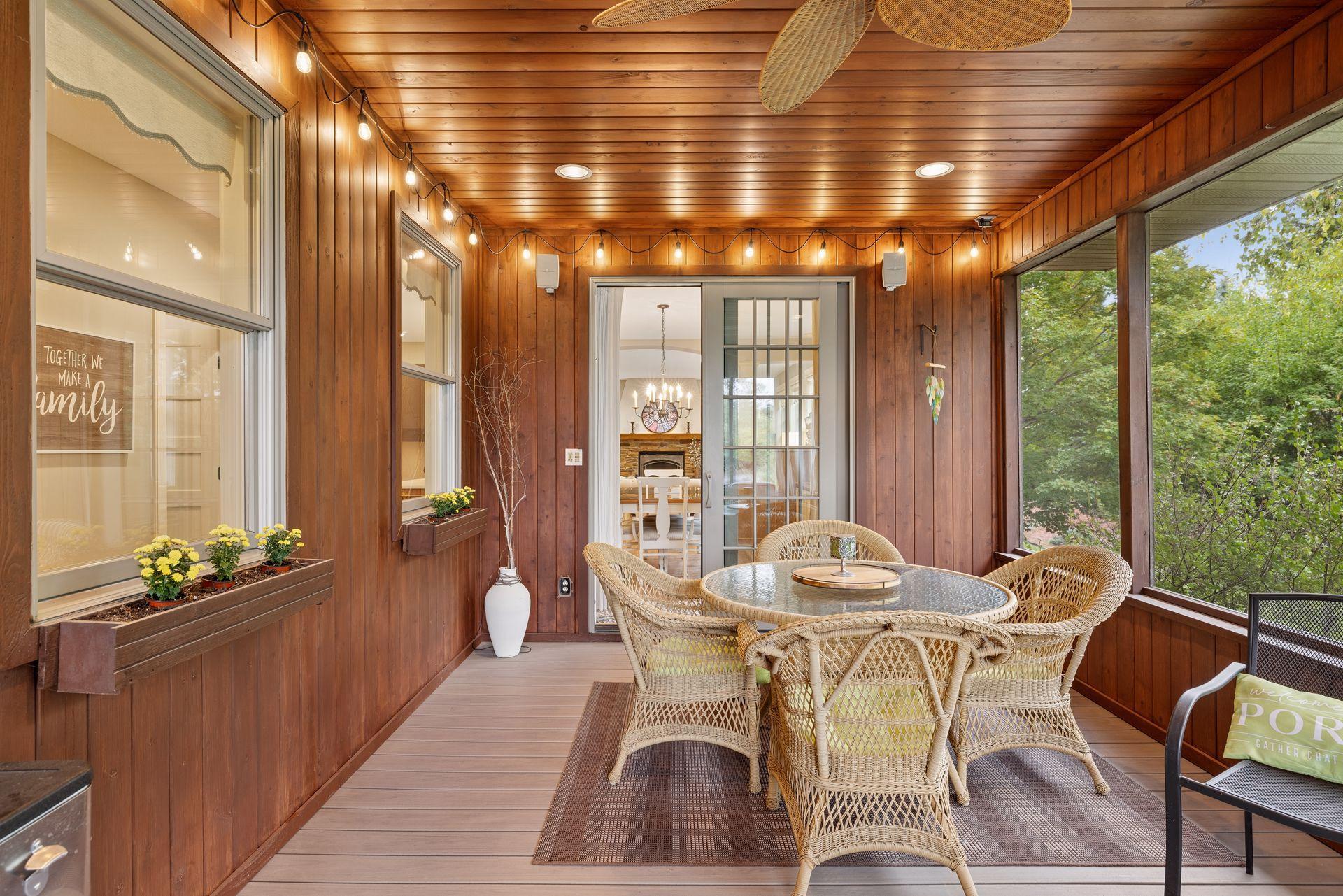 Wood-paneled enclosed porch with wicker dining set, string lights, and windows overlooking trees.
