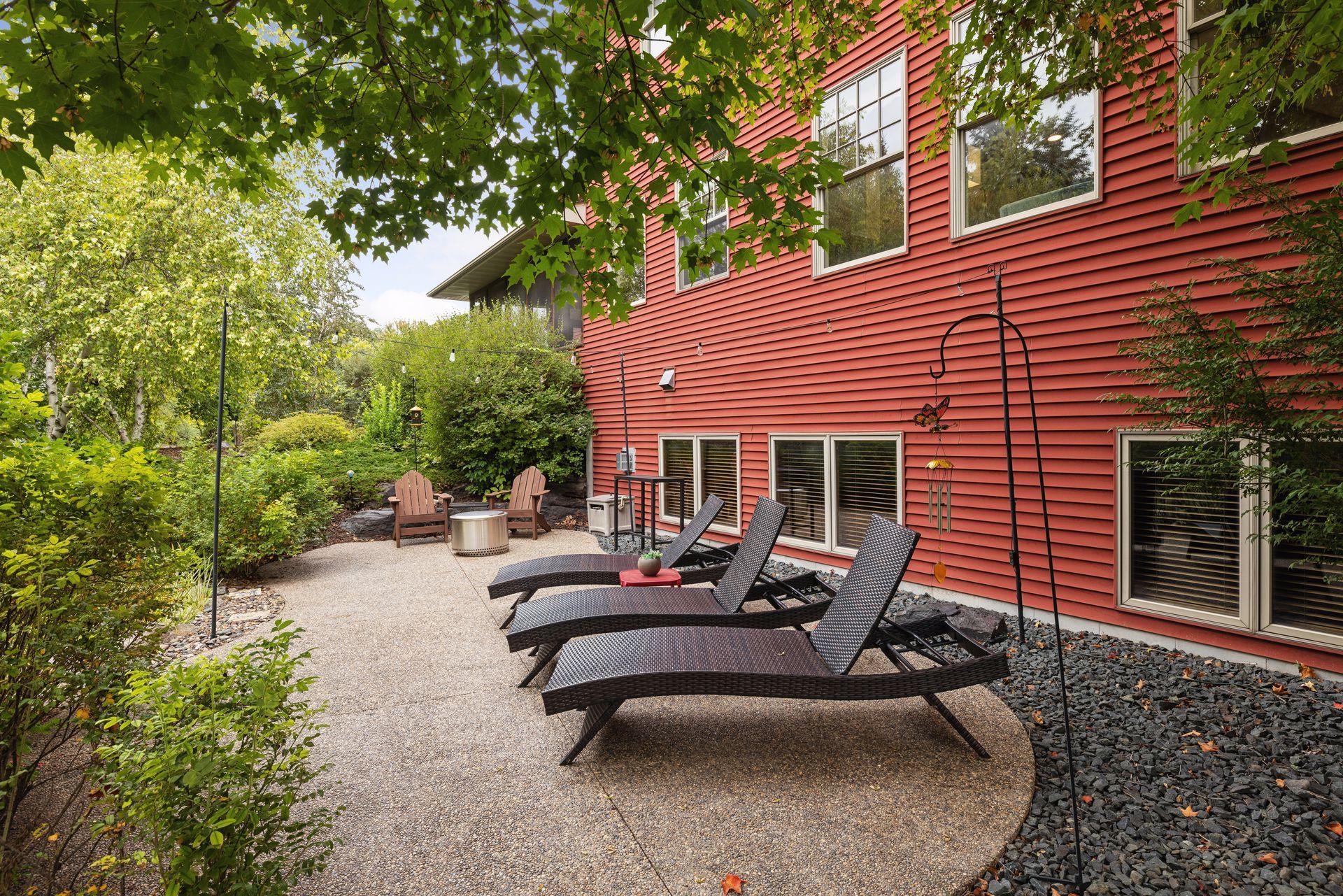 Patio beside a red-sided house with lounge chairs, a fire pit, and lush landscaping.