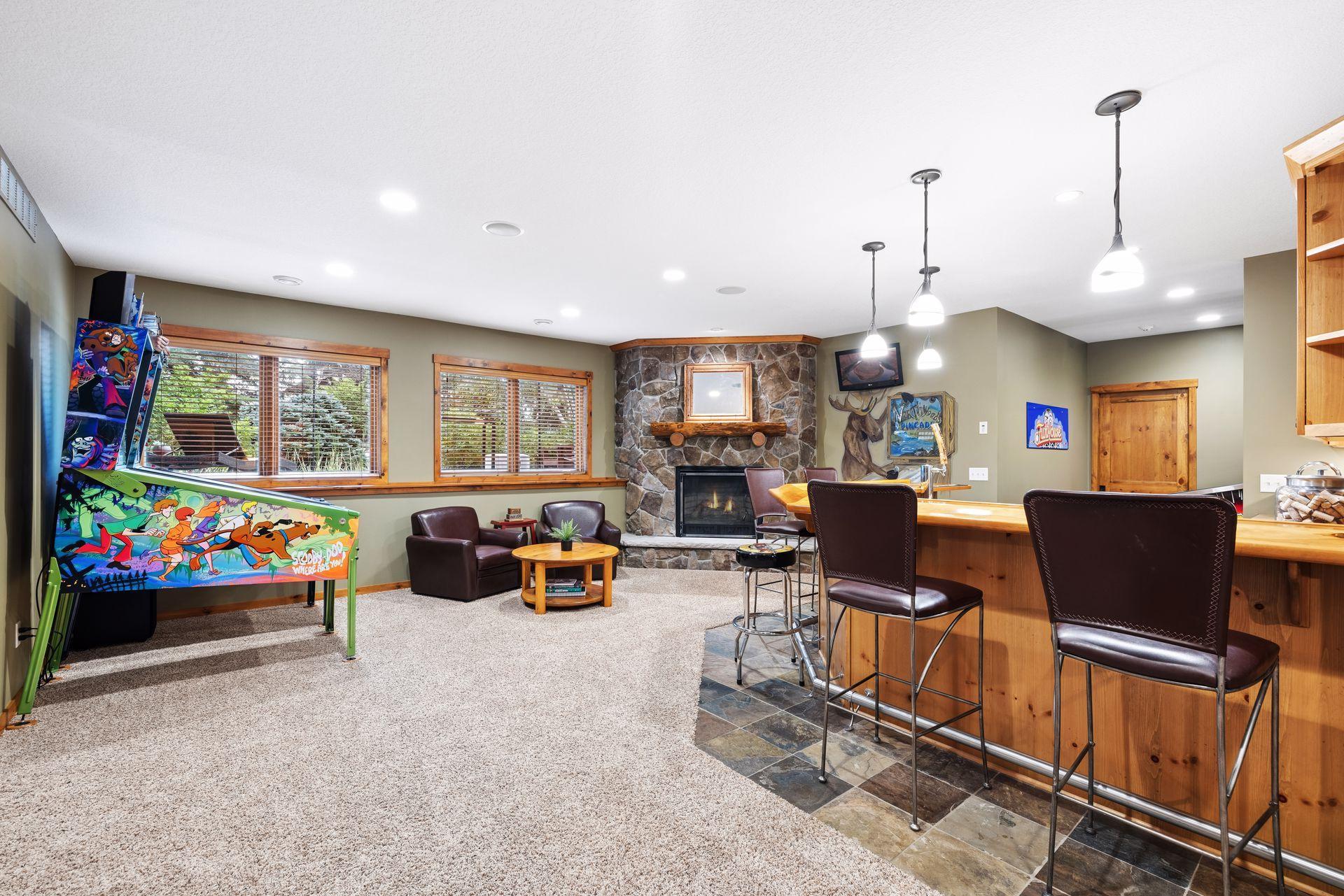 Furnished recreation room with a stone fireplace, wood bar, leather chairs, and colorful pinball machine.