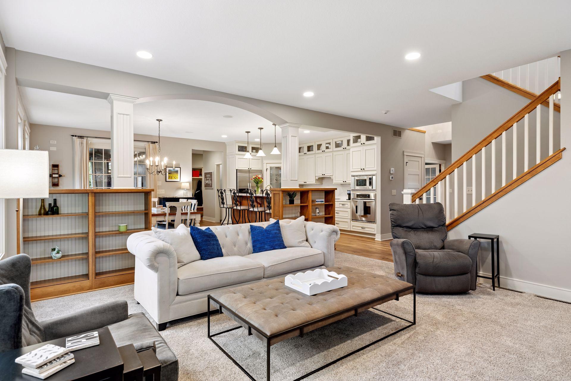 Open-plan living room with a white sofa, adjoining white kitchen, and staircase with wood handrail.