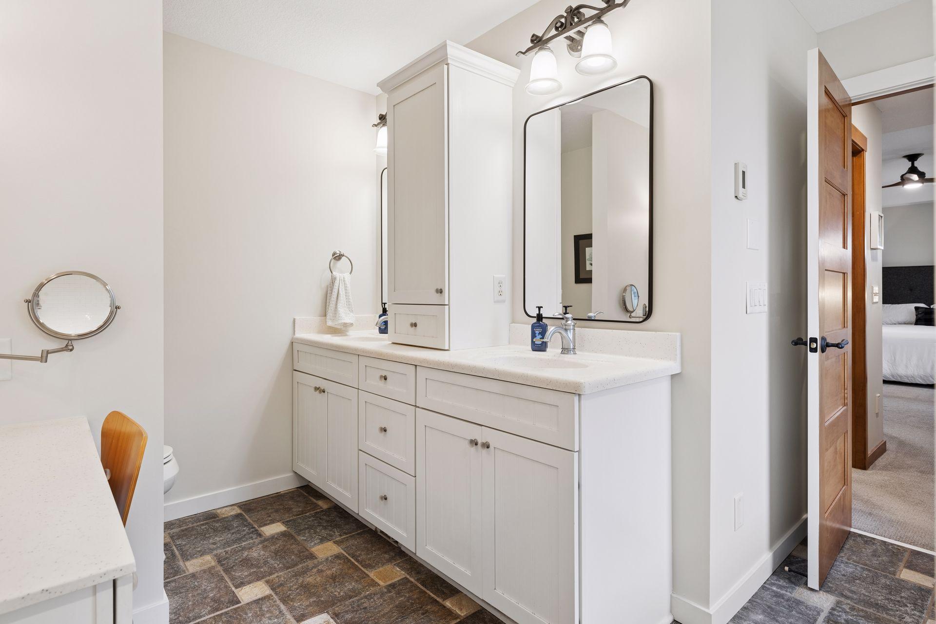 Bathroom vanity with white cabinetry, double sinks, a large mirror, and dark tile flooring.