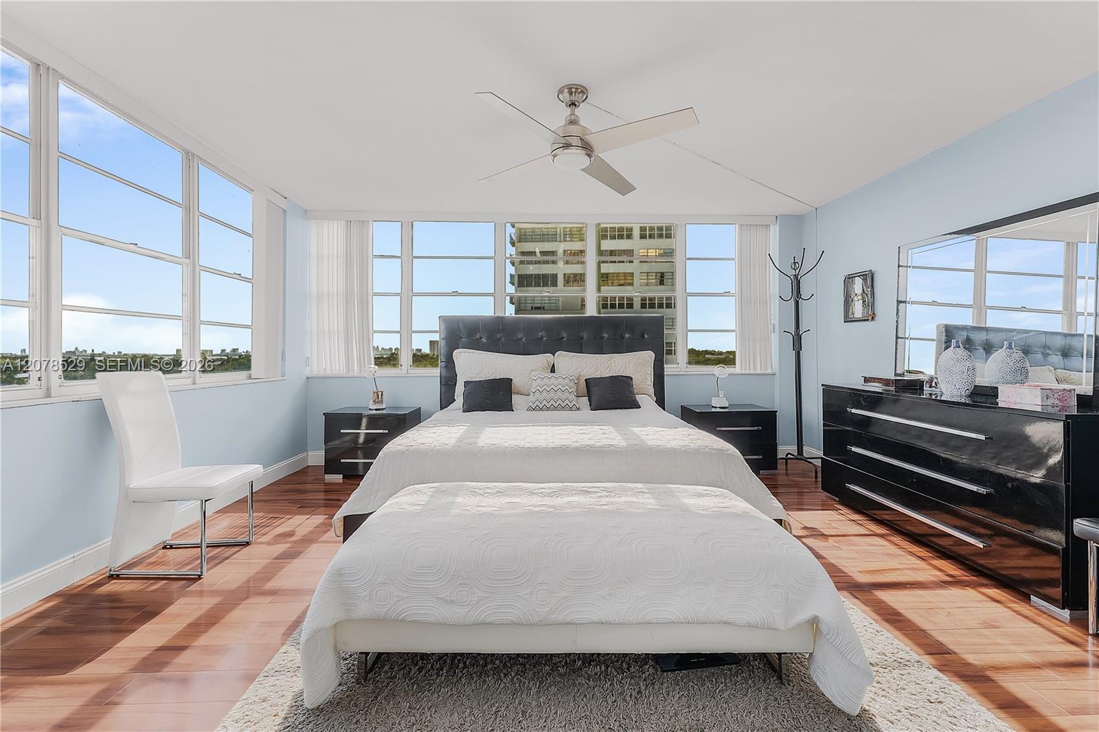 Staged bedroom with a large bed, corner windows, glossy wood floors, and a pale blue wall.