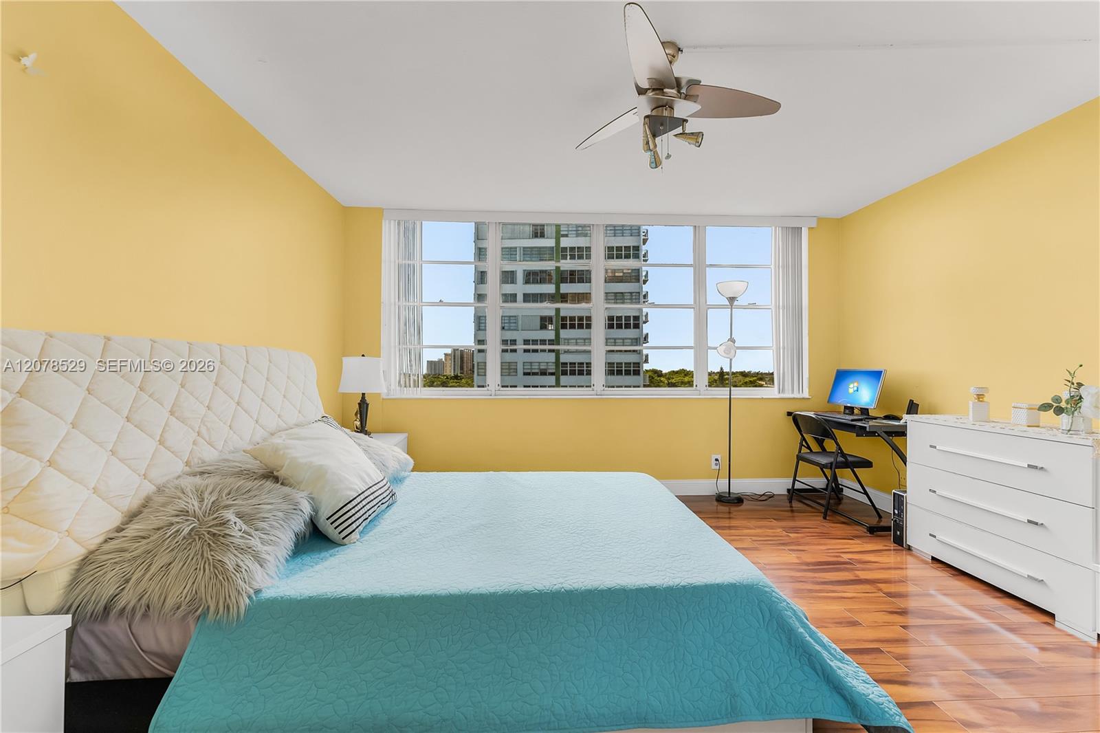 Bedroom with yellow walls, a turquoise bedspread, hardwood floors, and windows facing a nearby high-rise.