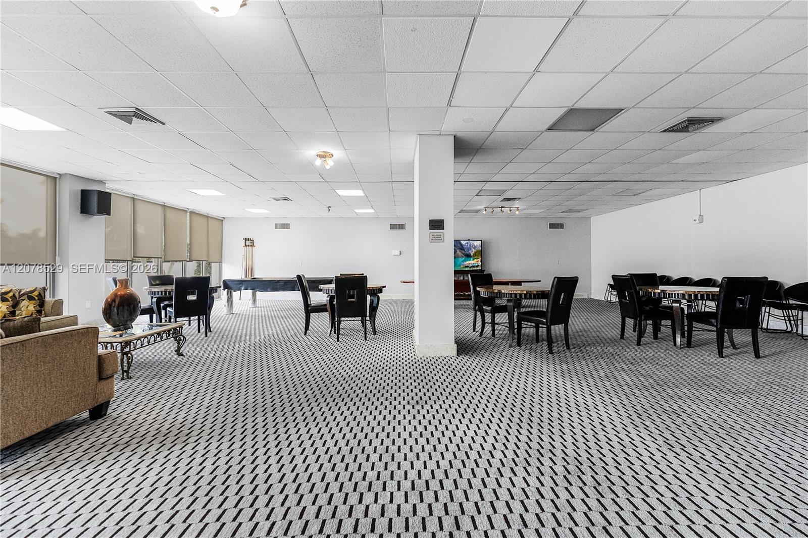 Furnished common room with patterned carpet, dining tables, chairs, sofa, and television.