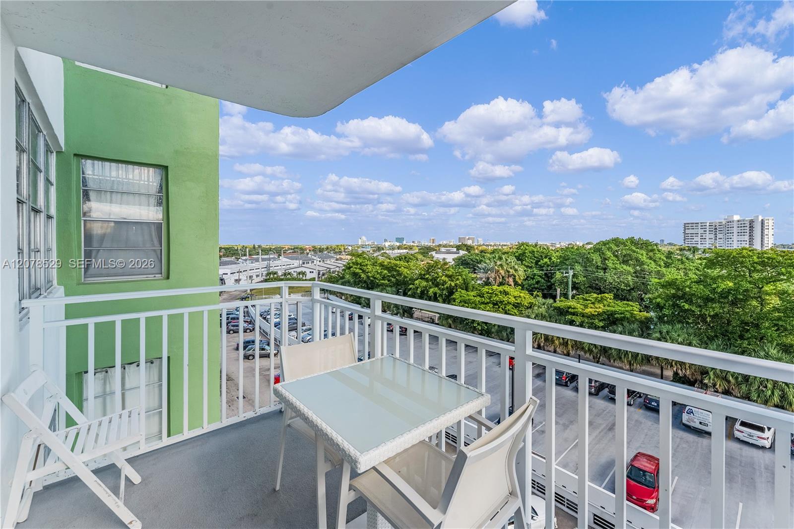 Apartment balcony with patio table and chairs overlooking trees, buildings, and a parking lot.