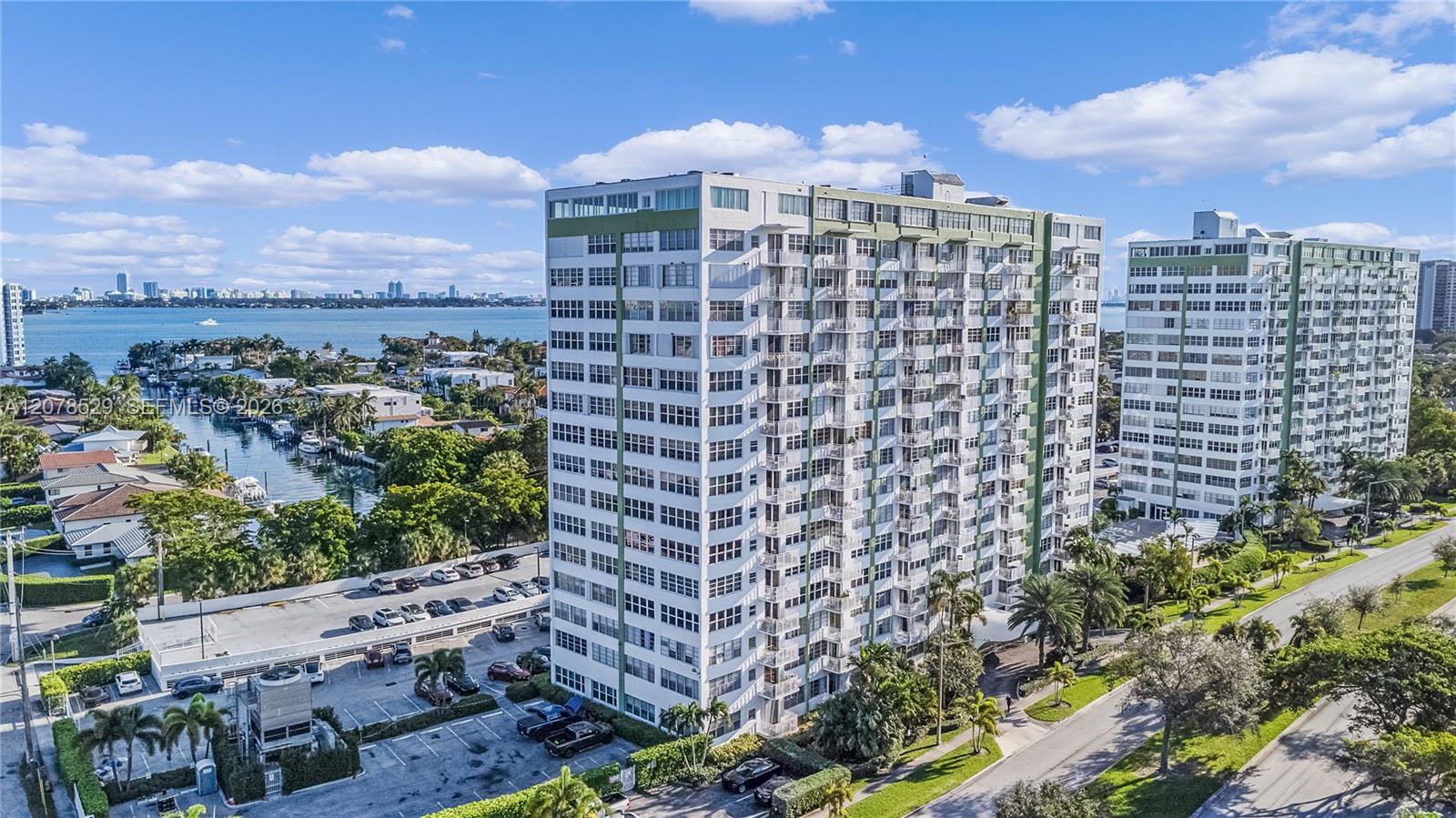 Aerial view of waterfront condominium towers beside a canal, bay, and distant city skyline.