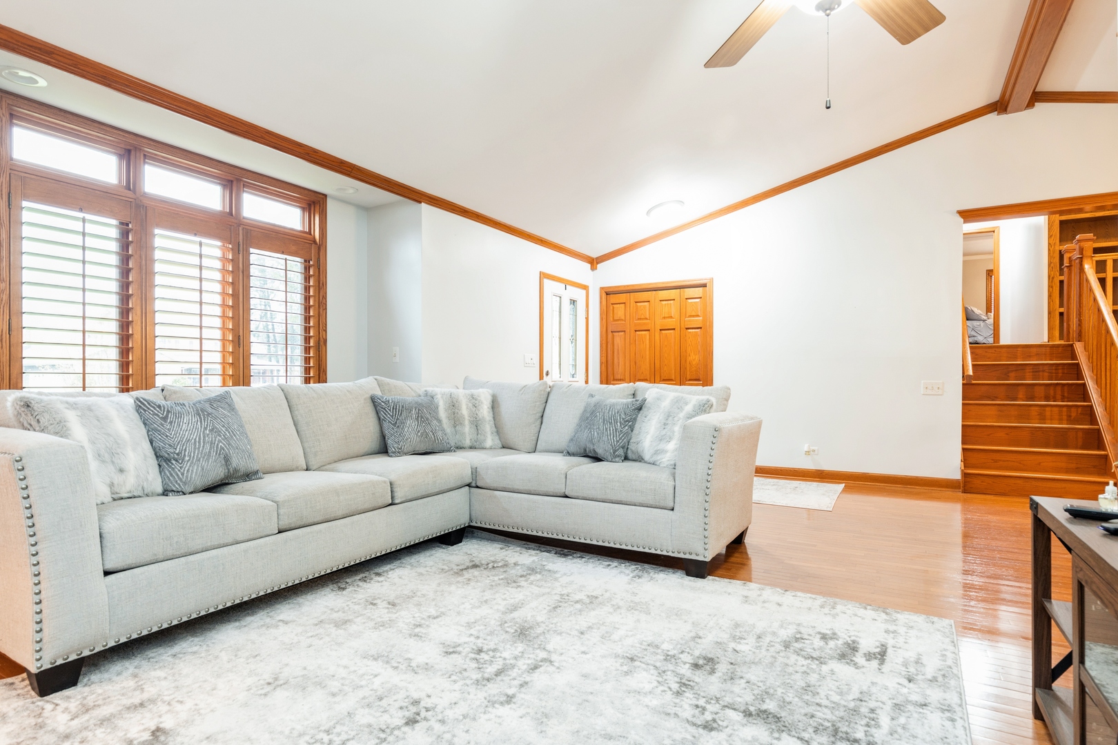 Living room with a gray sectional, shuttered windows, hardwood floors, and a wooden staircase.