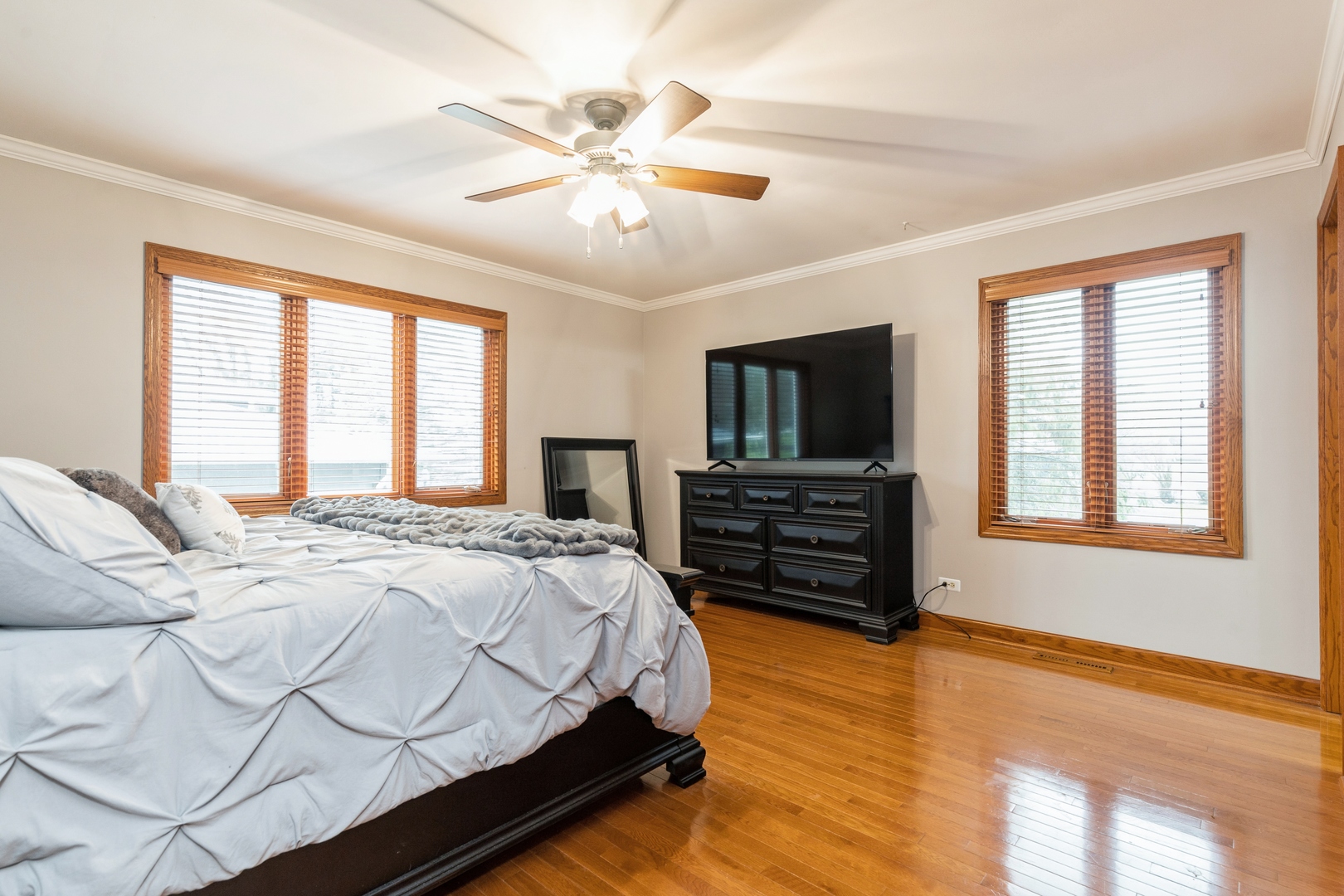 Furnished bedroom with a large bed, glossy hardwood floors, wood-trimmed windows, and a ceiling fan.