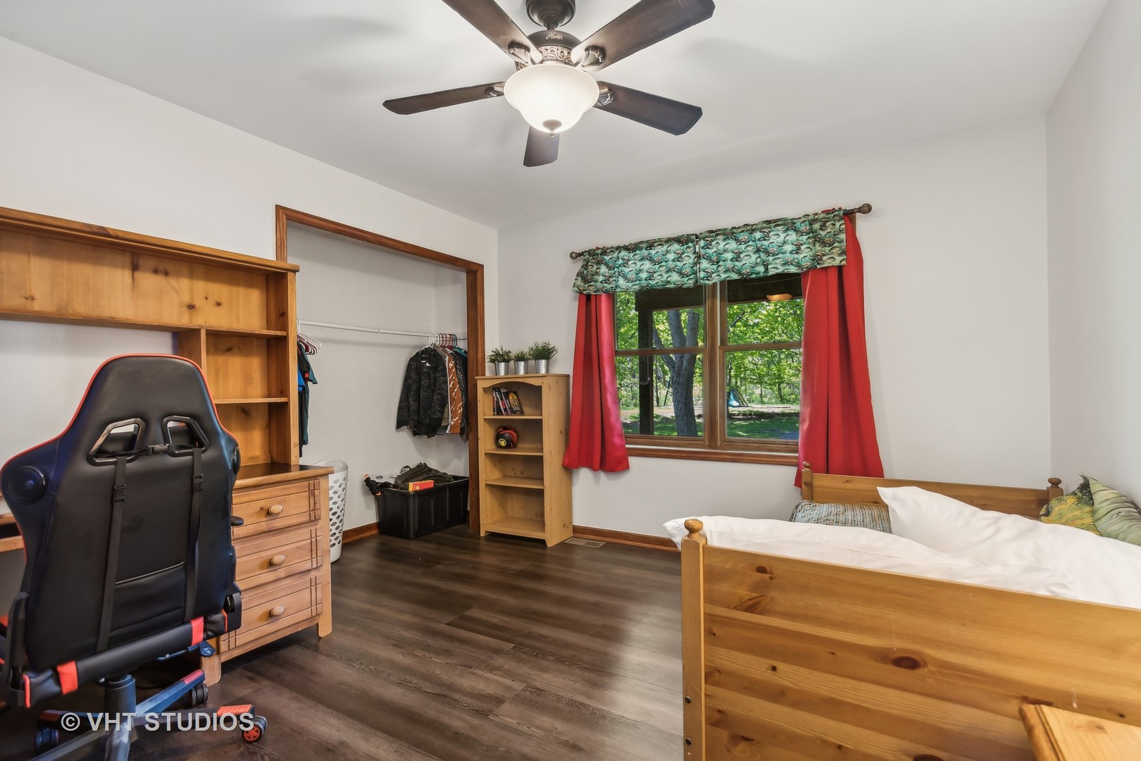Bedroom with a wooden bed, gaming chair, open closet, and red curtains framing a window.