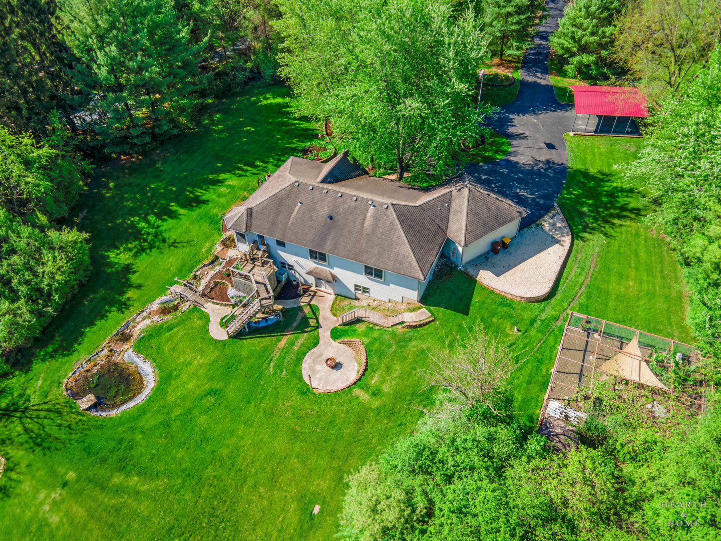 Aerial view of a house amid expansive lawns and trees, with patios, a pond, and a covered driveway structure.