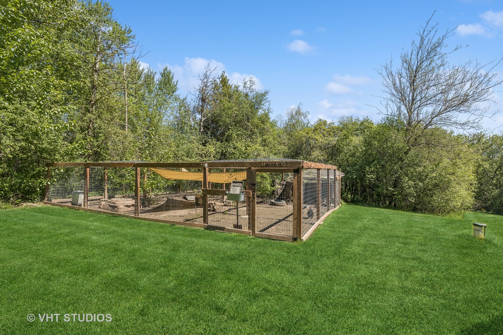 Wooden animal enclosure with wire mesh fencing on a spacious lawn surrounded by trees.