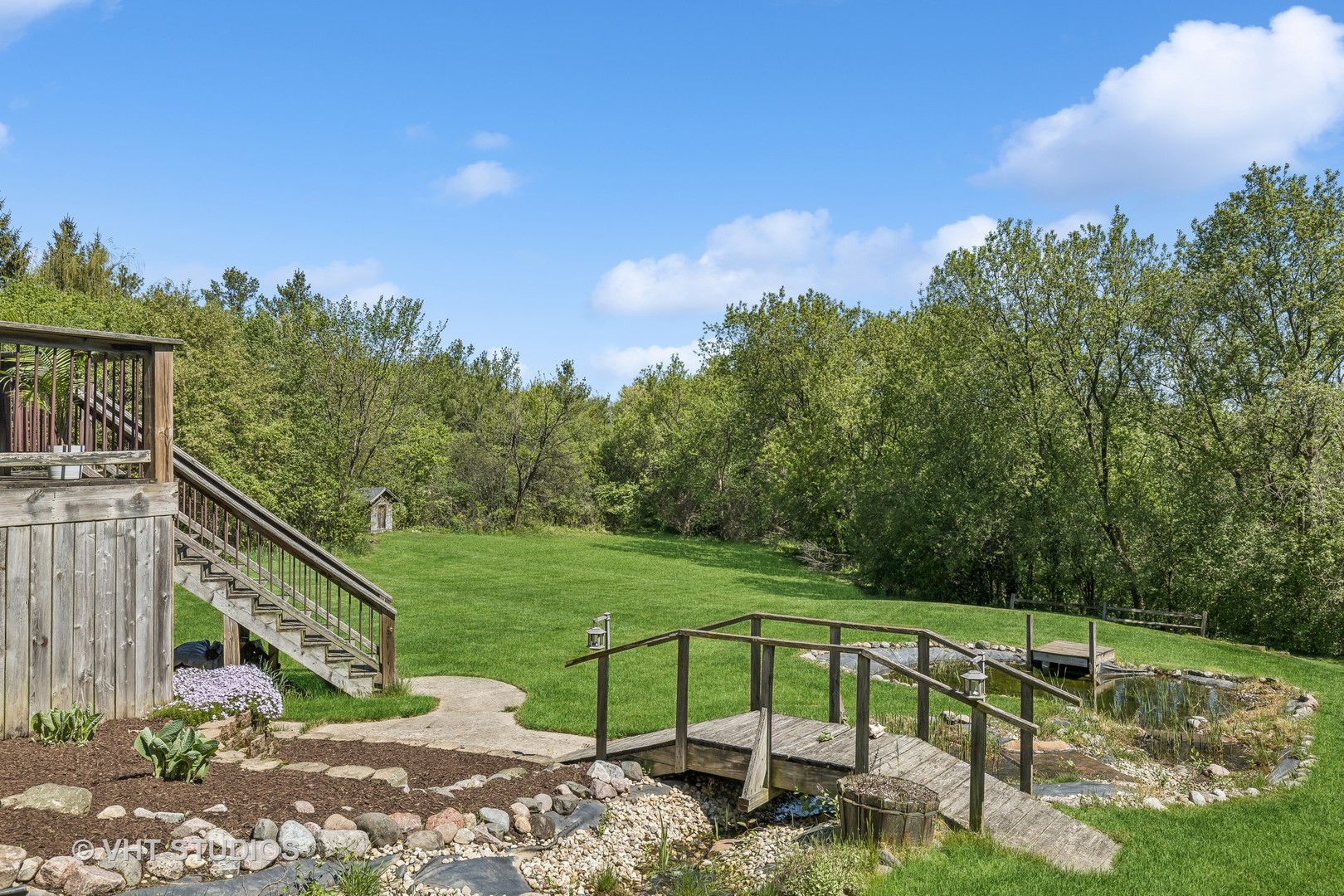 Backyard with a wooden footbridge over a rock-edged pond, lawn, mature trees, and deck stairs.