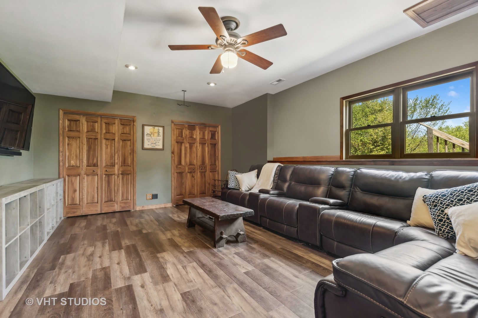 Living room with a dark leather sectional, wood-look floors, and windows overlooking trees.