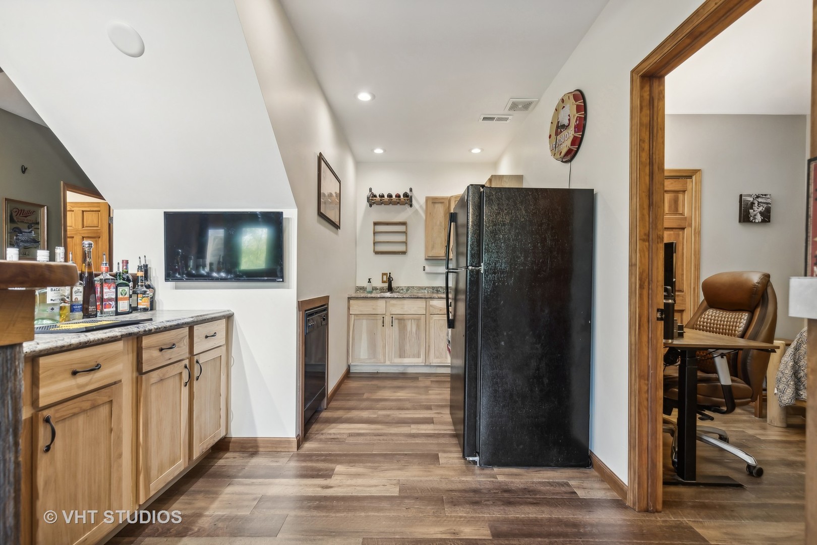 Basement kitchenette with wood cabinets, black refrigerator, and an adjacent office doorway.