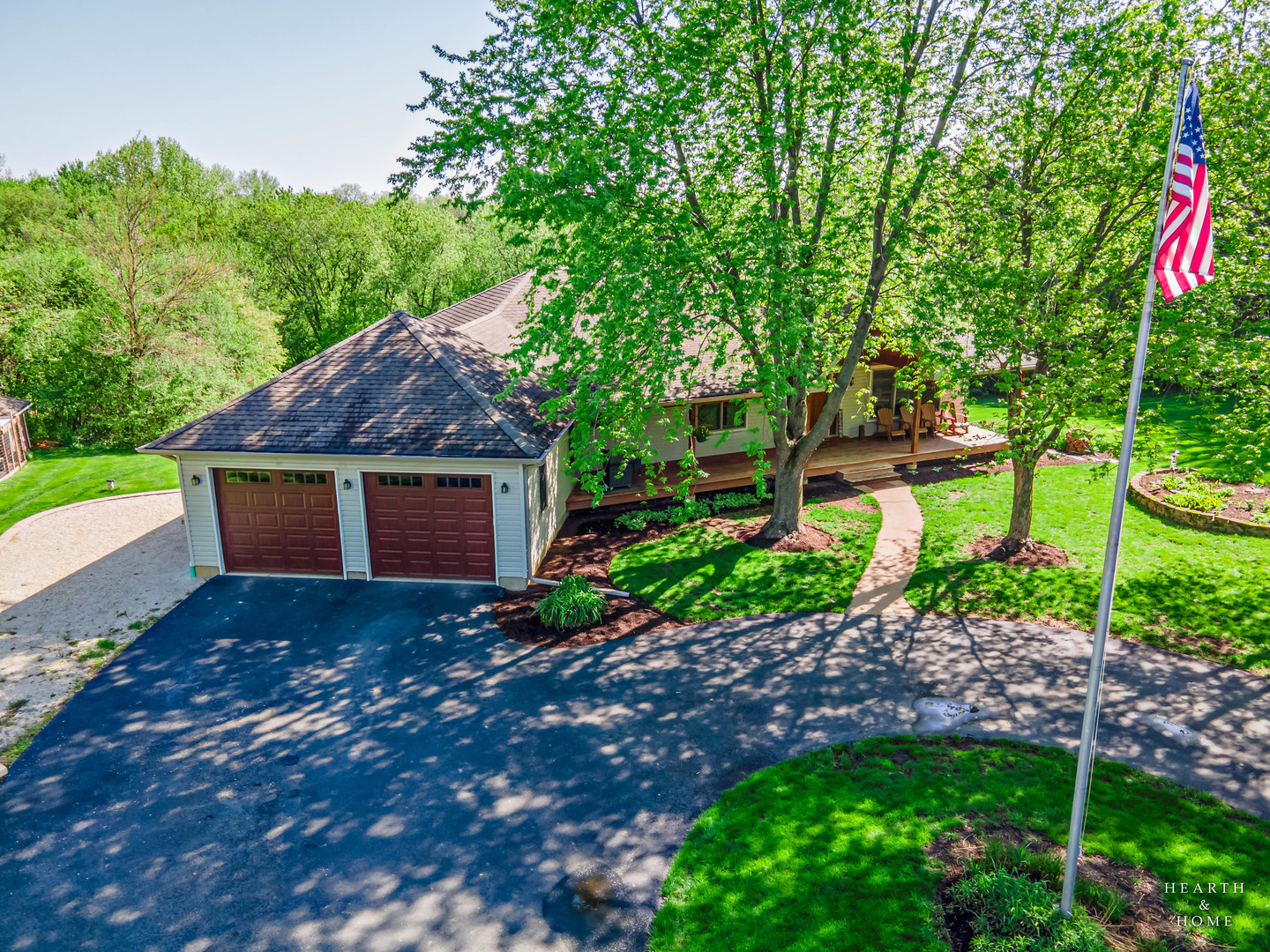 House with a two-car garage, shaded driveway, covered porch, mature trees, and an American flag.