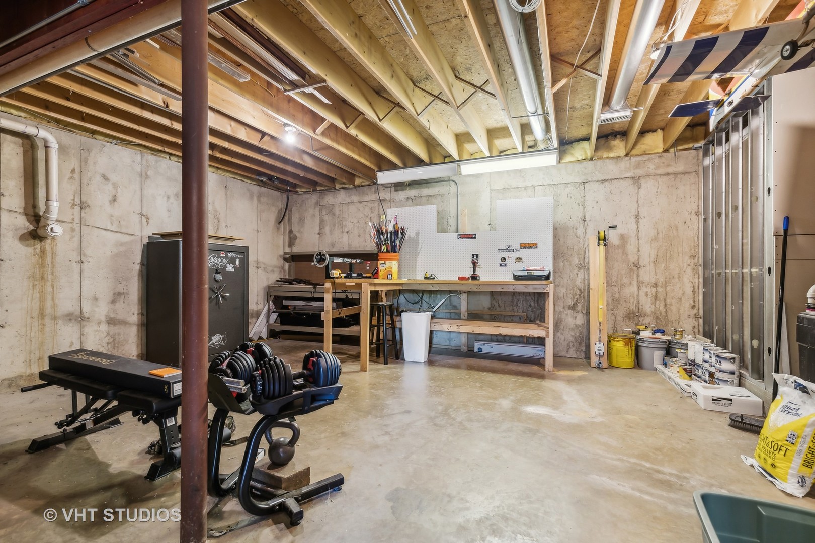 Unfinished basement workshop with a workbench, pegboard, weights, and exposed ceiling joists.
