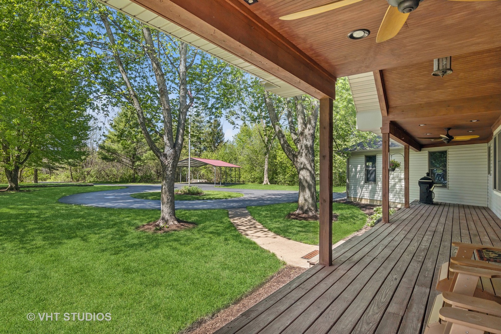 Covered wooden deck overlooking a lawn, mature trees, circular driveway, and red-roofed carport.