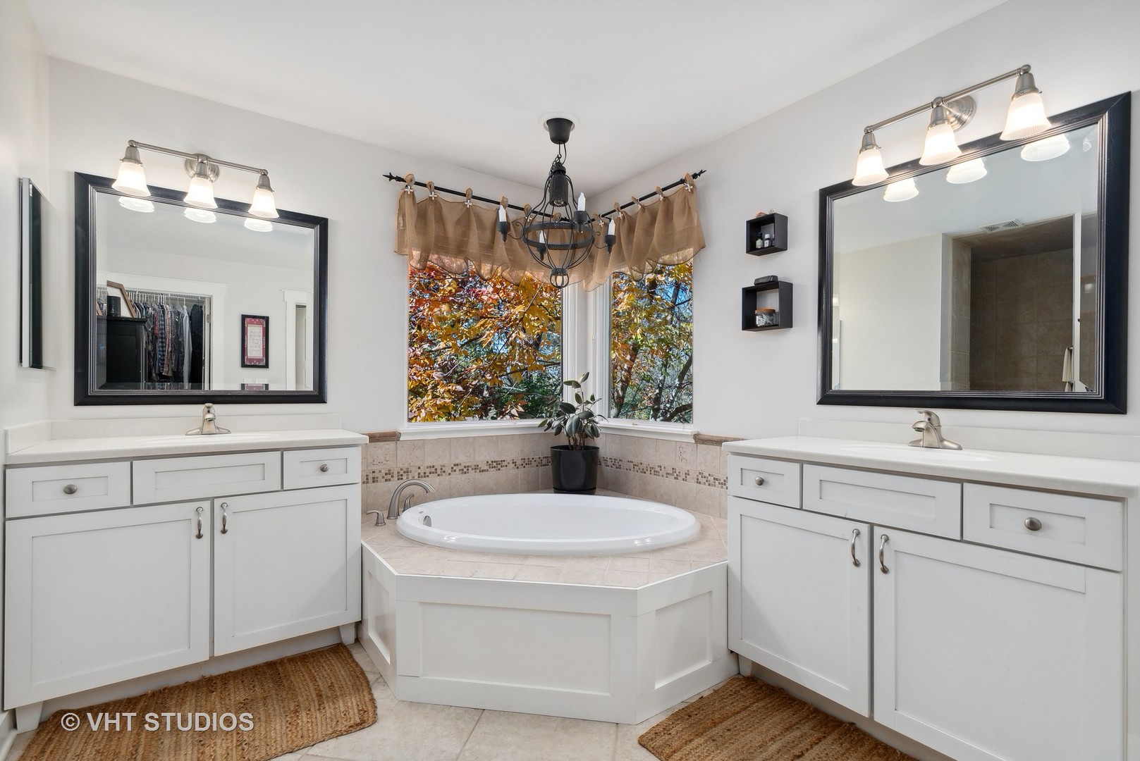 Bathroom with dual white vanities and a corner soaking tub beneath windows overlooking autumn foliage.