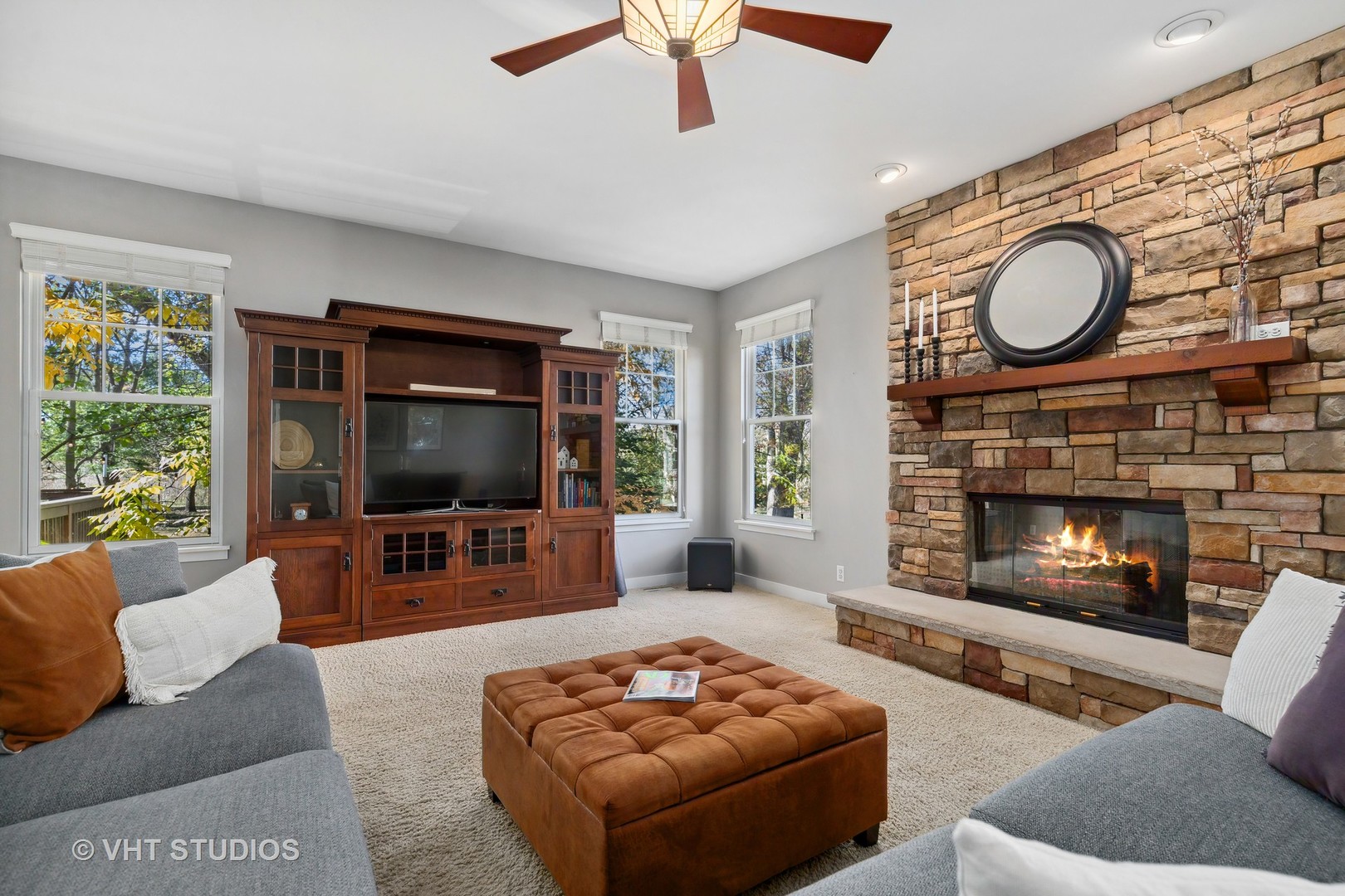 Living room with a stone fireplace, built-in entertainment cabinet, gray sofas, and brown tufted ottoman.