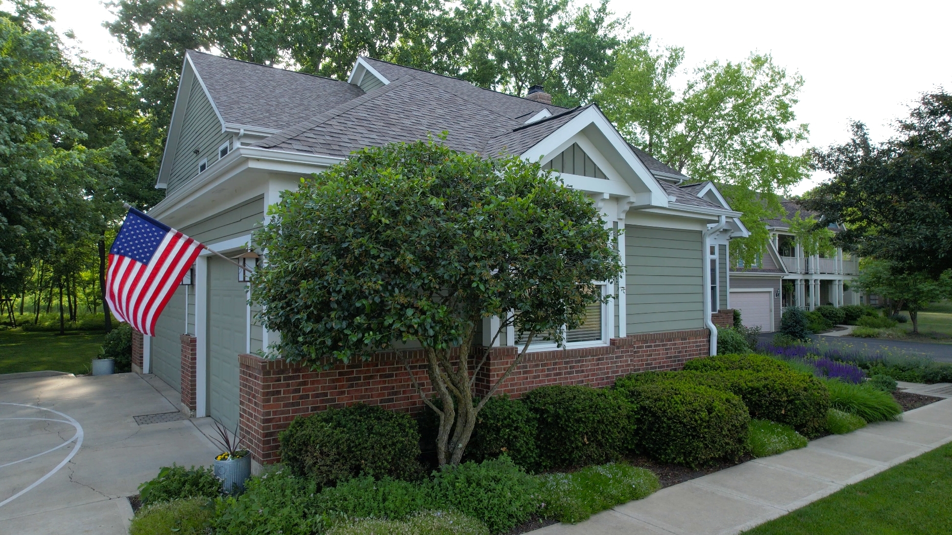 Gray-green house with an attached garage, brick foundation, landscaped shrubs, and an American flag.