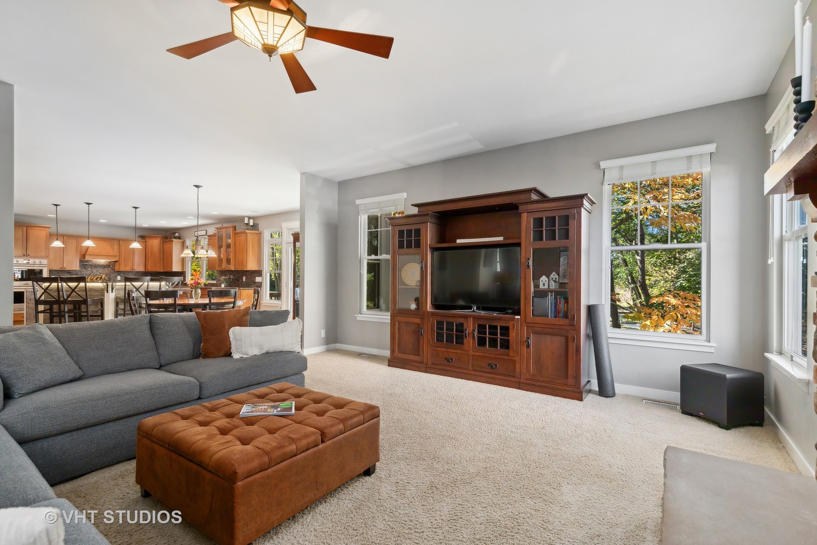 Living room with gray sectional, leather ottoman, and wooden entertainment center beside an open kitchen.