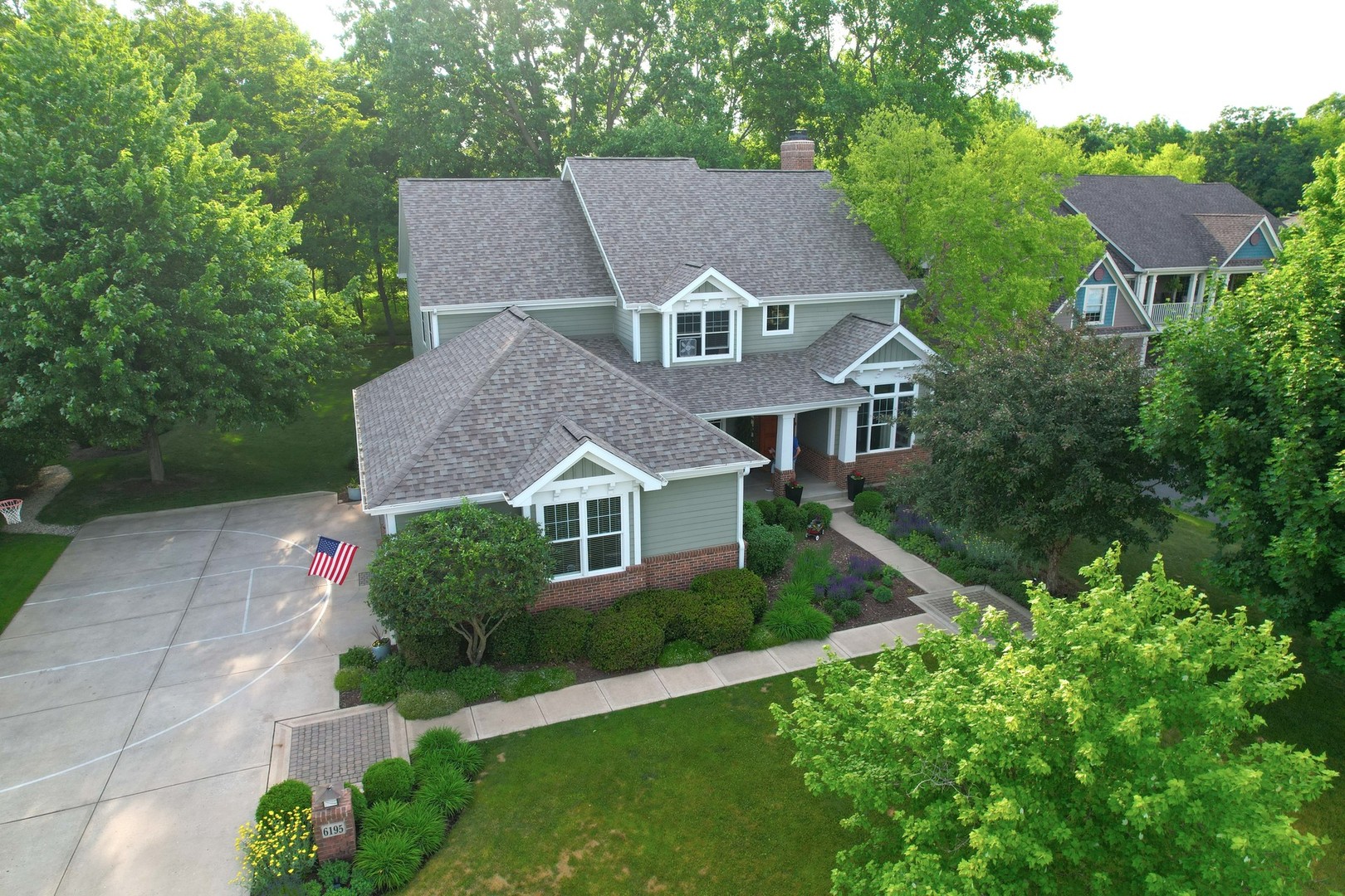 Aerial view of a green-sided home with a brick facade, landscaped grounds, and a basketball-marked driveway.
