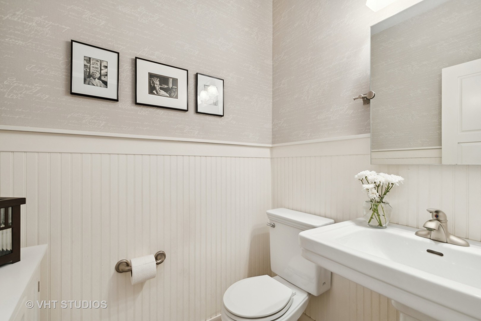 Half bathroom with a pedestal sink, white beadboard wainscoting, patterned wallpaper, and framed prints.