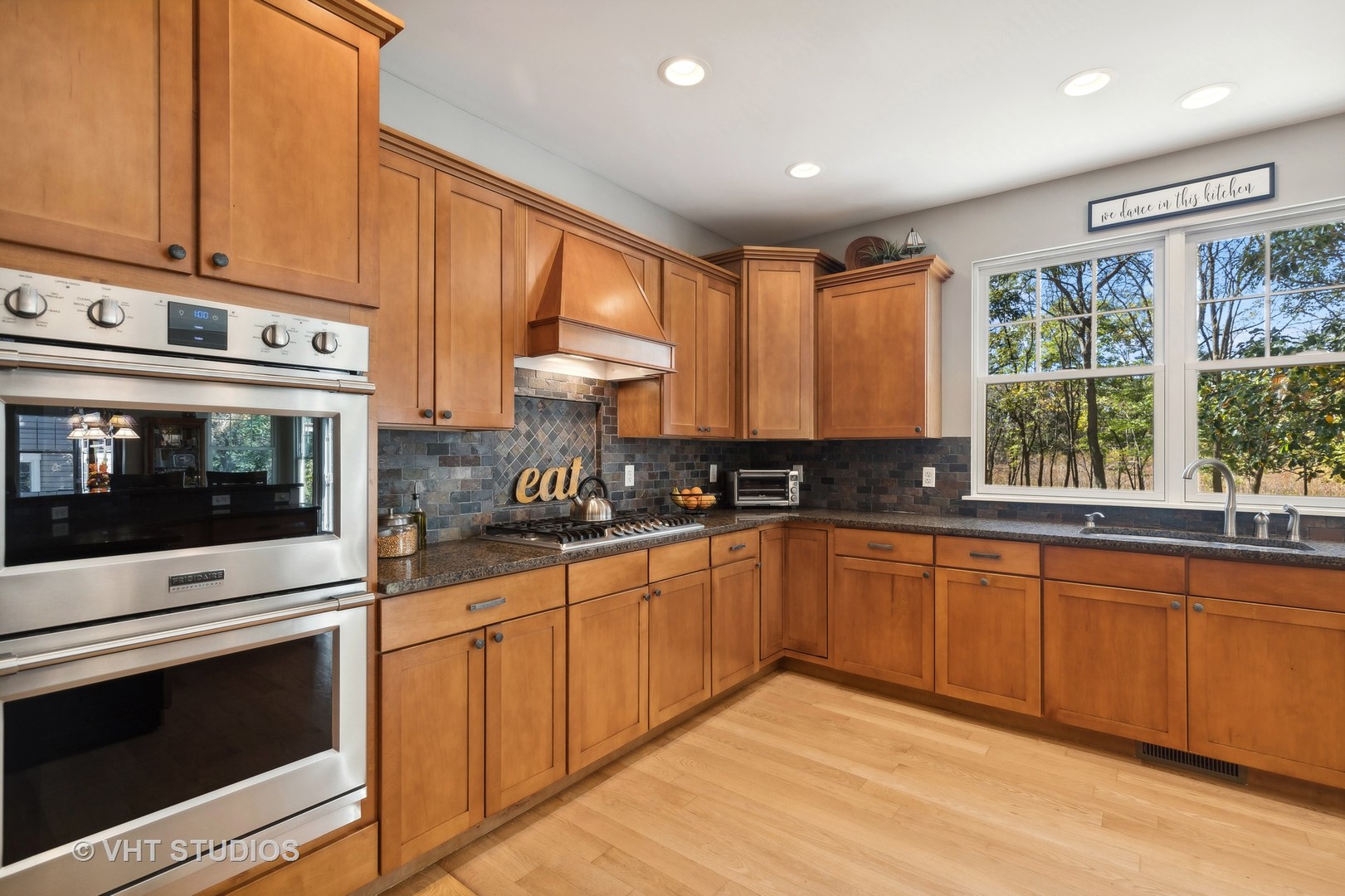 Kitchen with honey-toned wood cabinets, stainless double ovens, gas cooktop, and a large window overlooking trees.