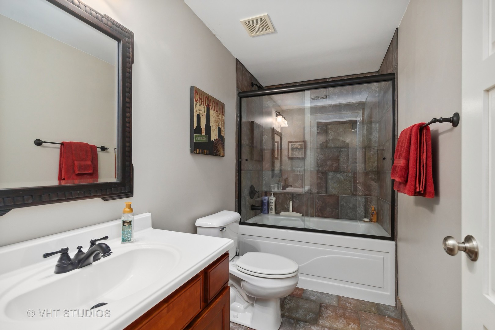 Bathroom with a white vanity, glass-enclosed tub, and mottled stone-look tile surround.
