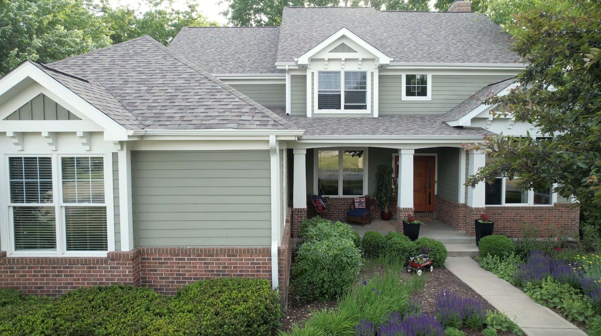 Two-story sage-green home with a covered porch, brick facade, and purple-flowered landscaping.