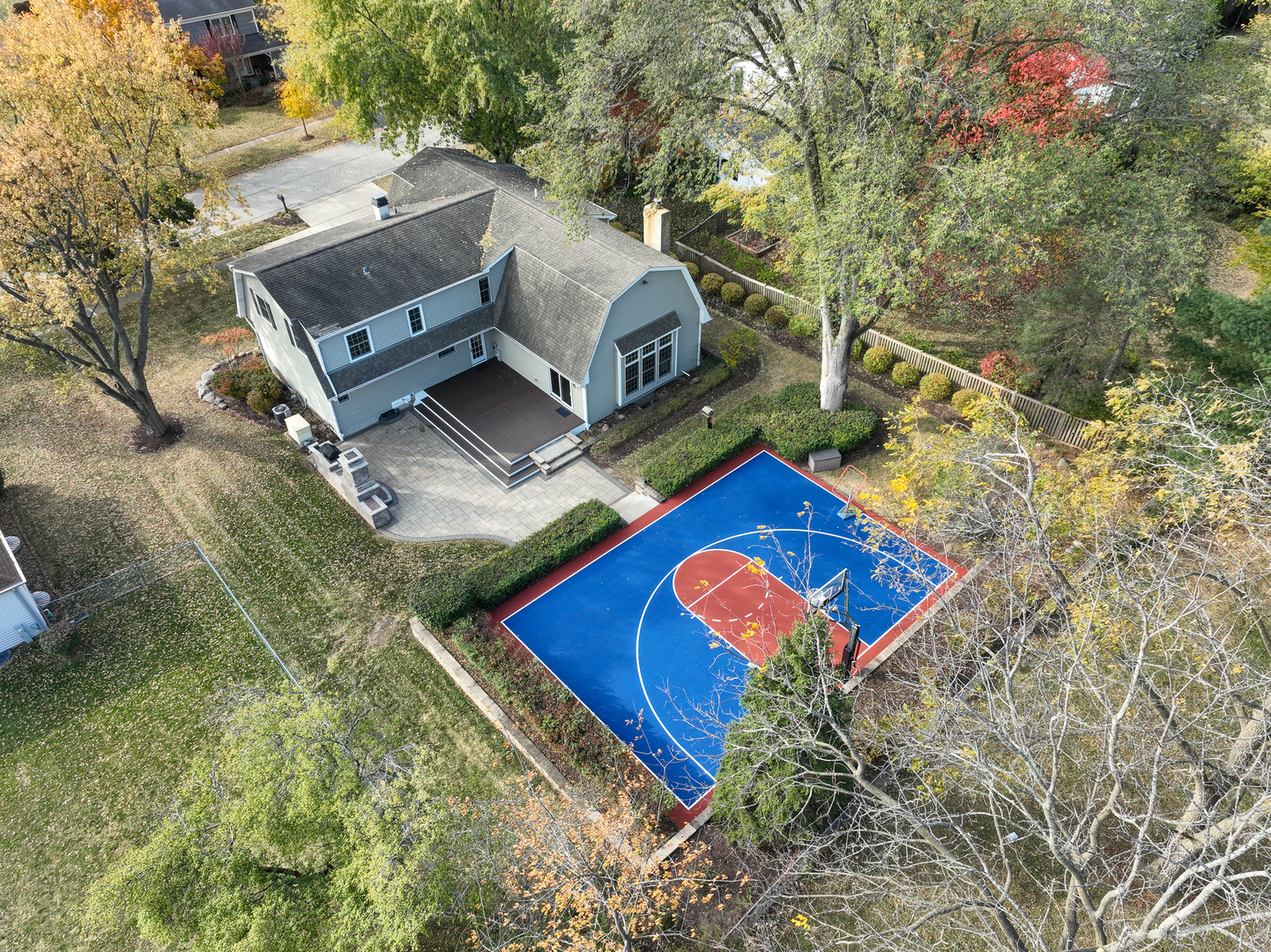 Aerial view of a gray home with a backyard basketball court and deck among autumn trees.