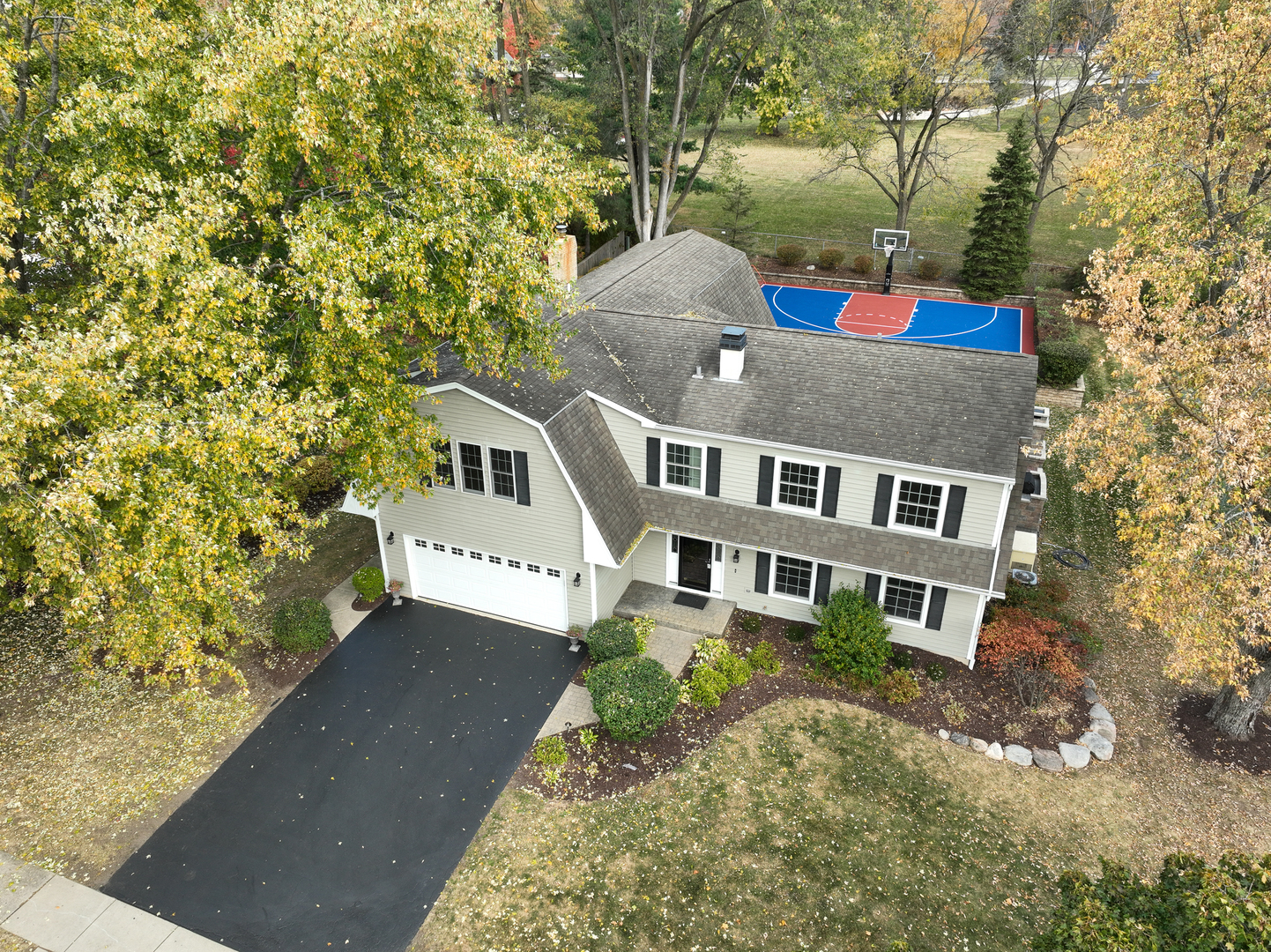 Aerial view of a two-story home with a backyard basketball court and mature autumn trees.