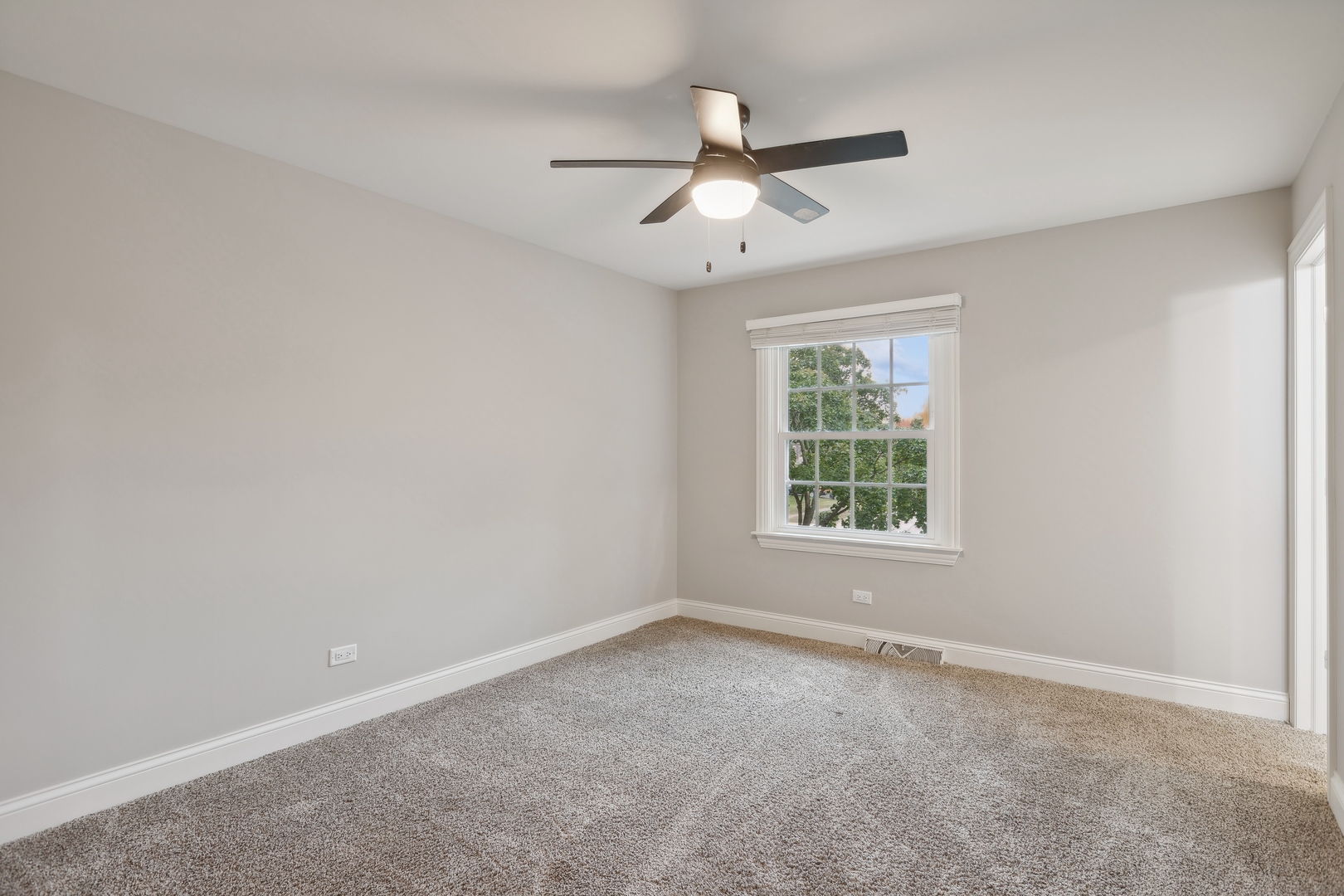Empty carpeted bedroom with a ceiling fan and a window overlooking trees.