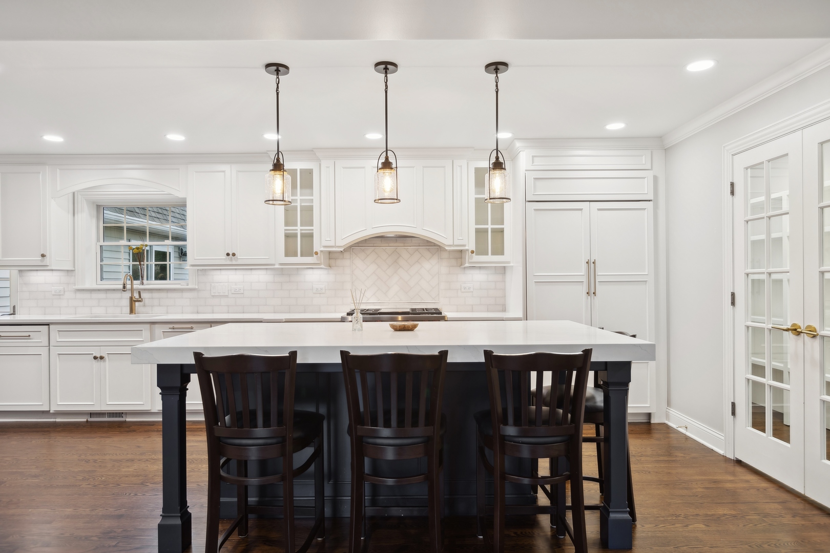 Bright white kitchen with a large island, dark bar stools, pendant lights, and hardwood floors.