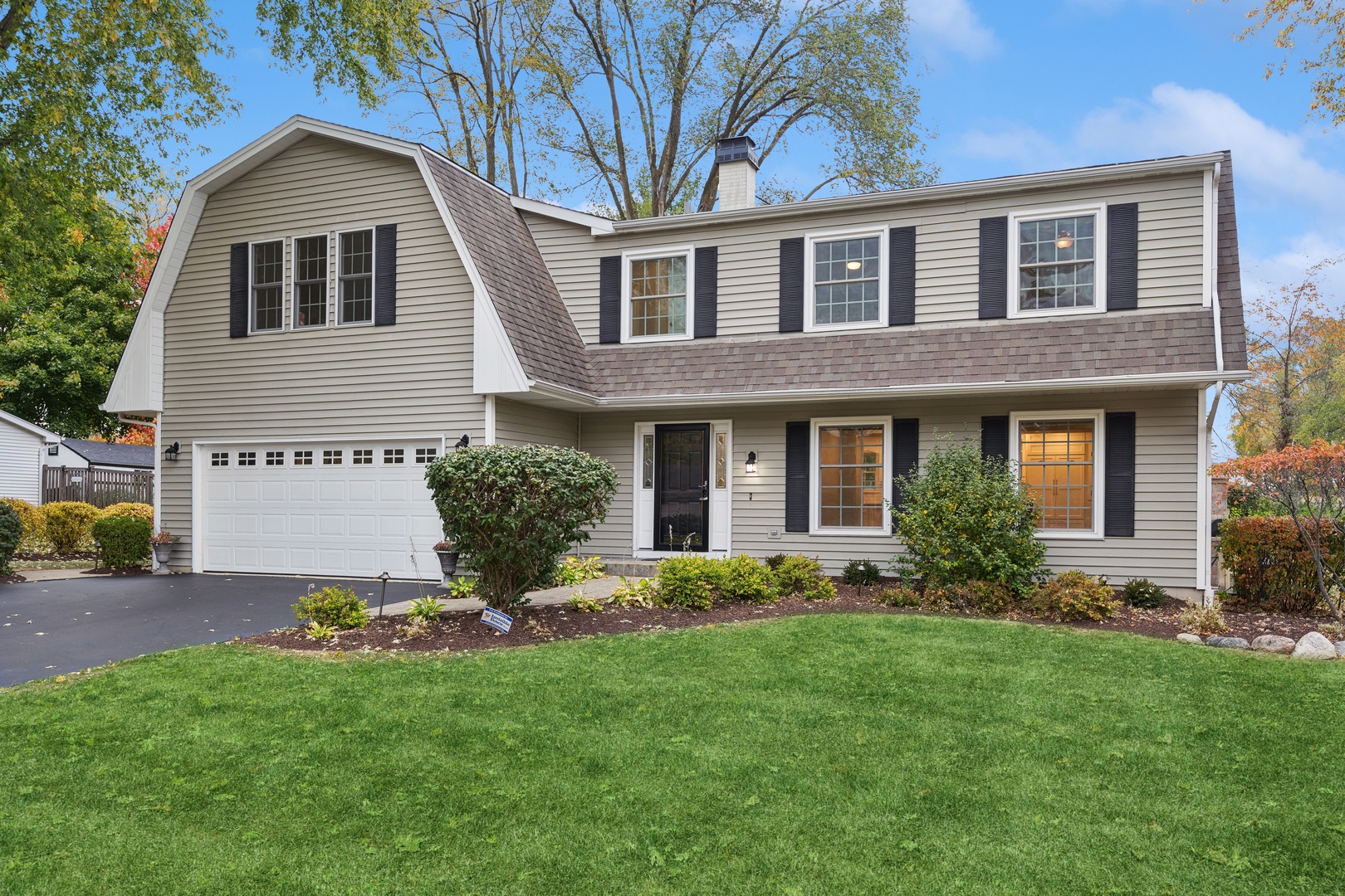 Two-story beige house with a gambrel roof, attached garage, black shutters, and landscaped lawn.