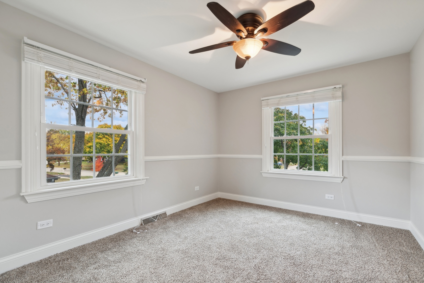 Empty bedroom with beige carpet, two windows, and a ceiling fan with light.