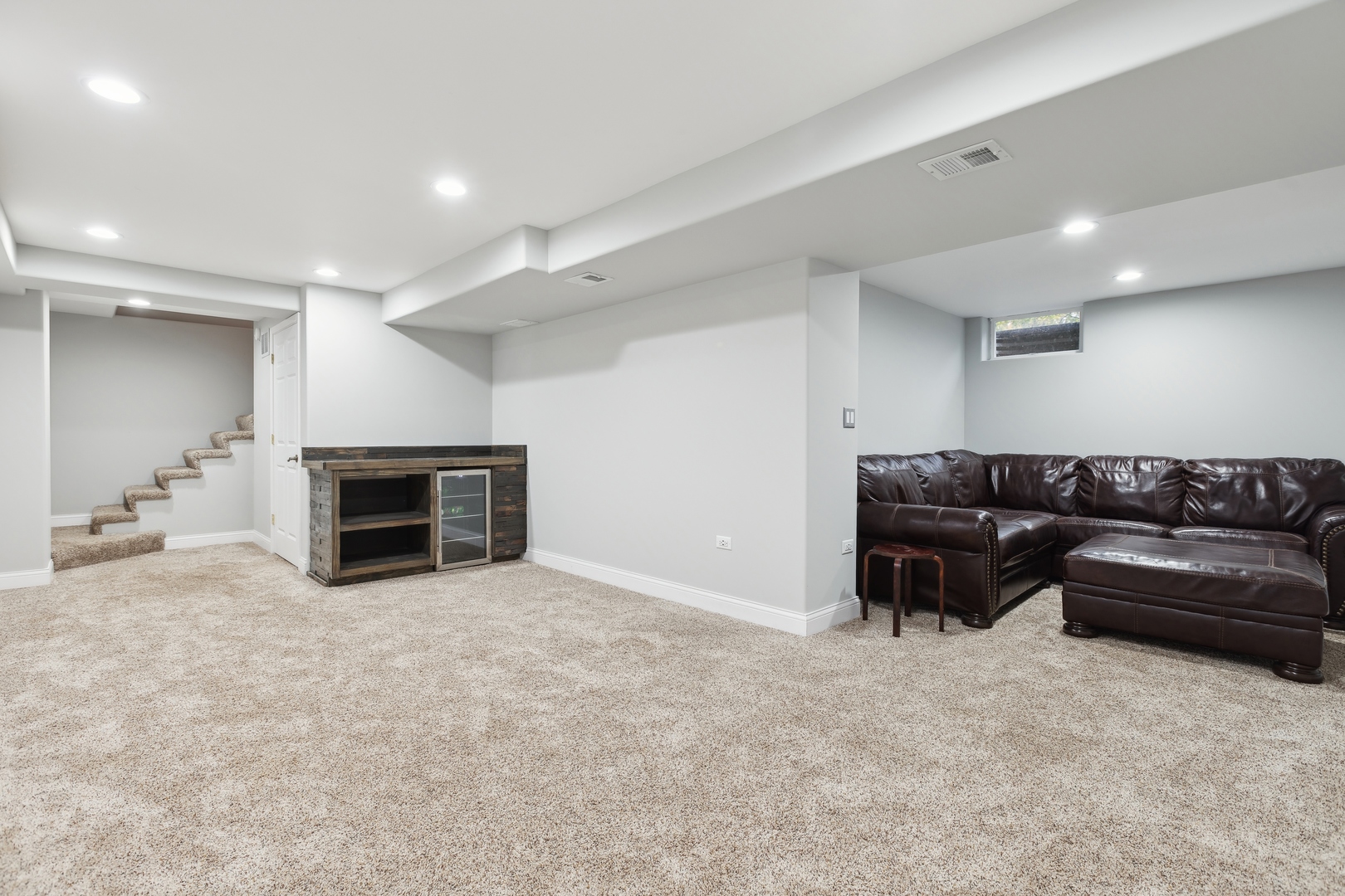 Finished basement family room with a leather sectional, beige carpet, beverage cabinet, and carpeted stairs.