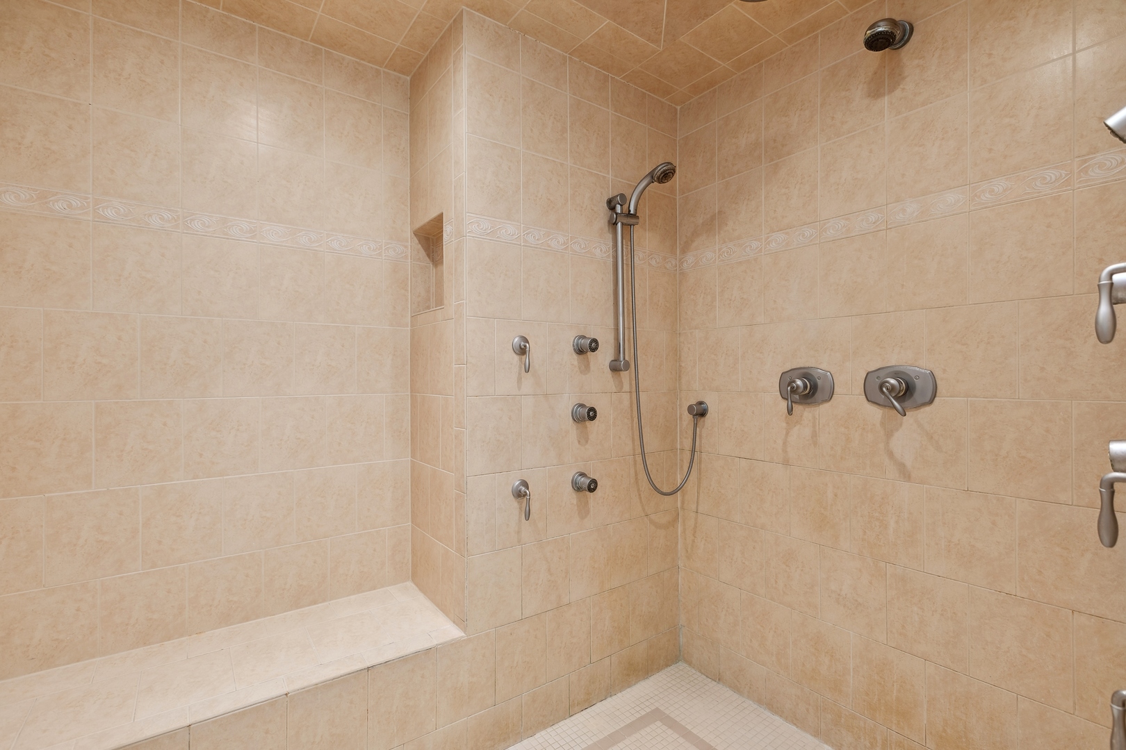 Beige-tiled shower with built-in bench, handheld showerhead, and multiple wall sprays.