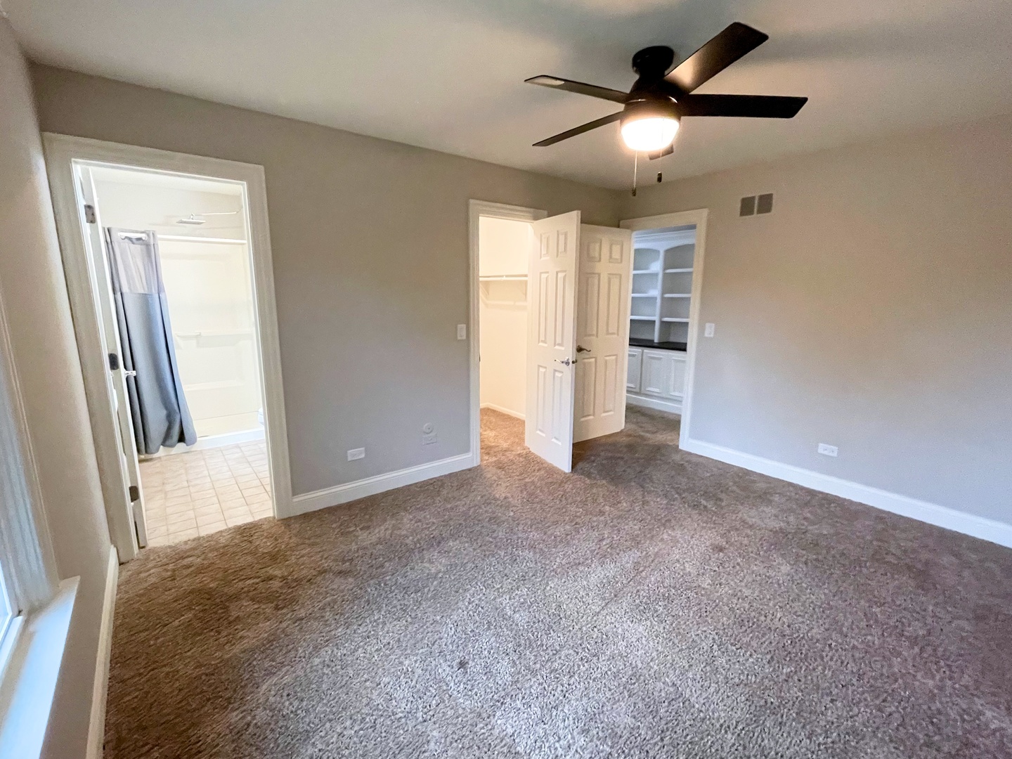 Empty bedroom with brown carpet, ceiling fan, and open doors to a bathroom and closet.
