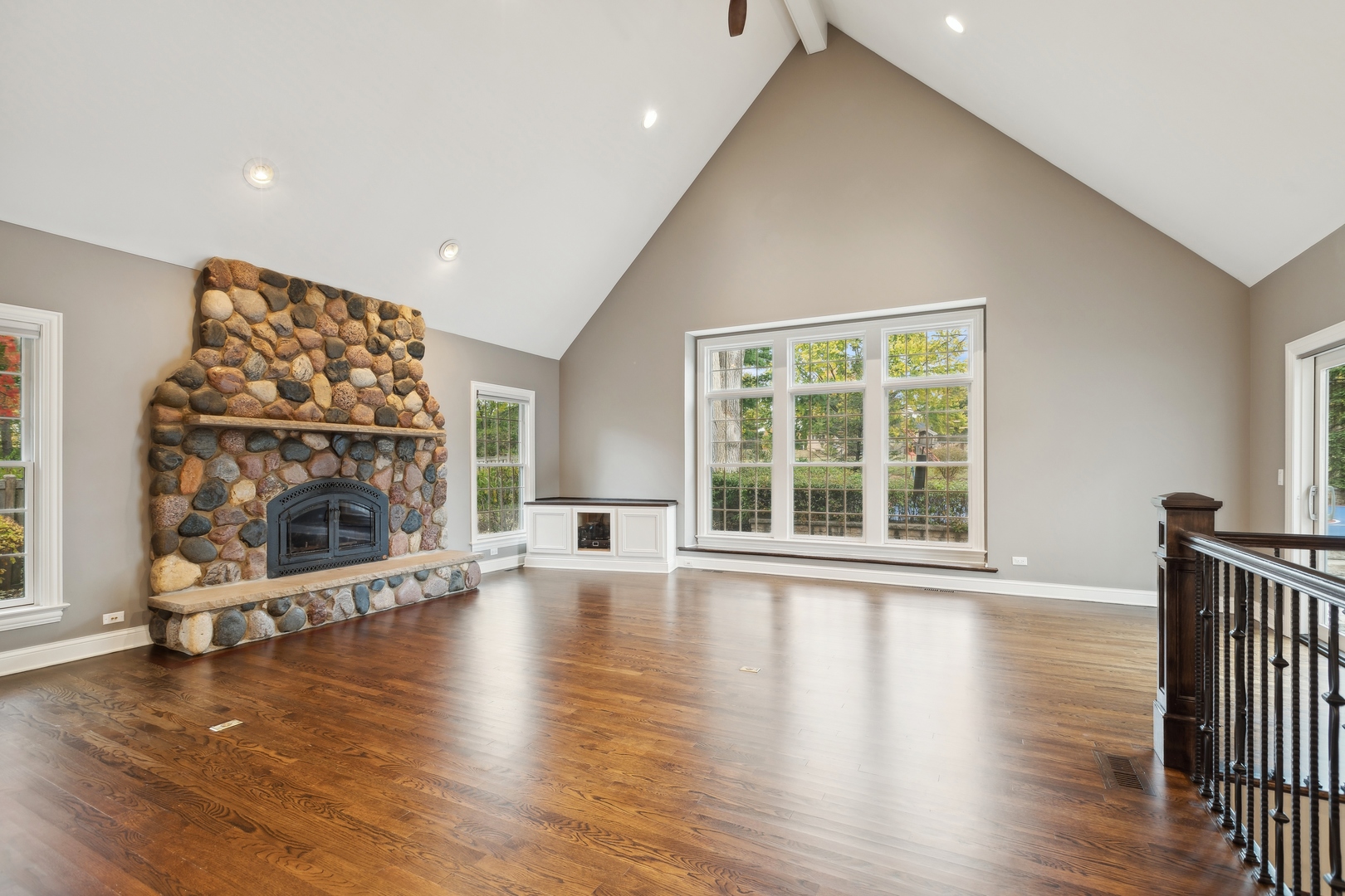 Living room with a vaulted ceiling, stone fireplace, hardwood floors, and large windows.