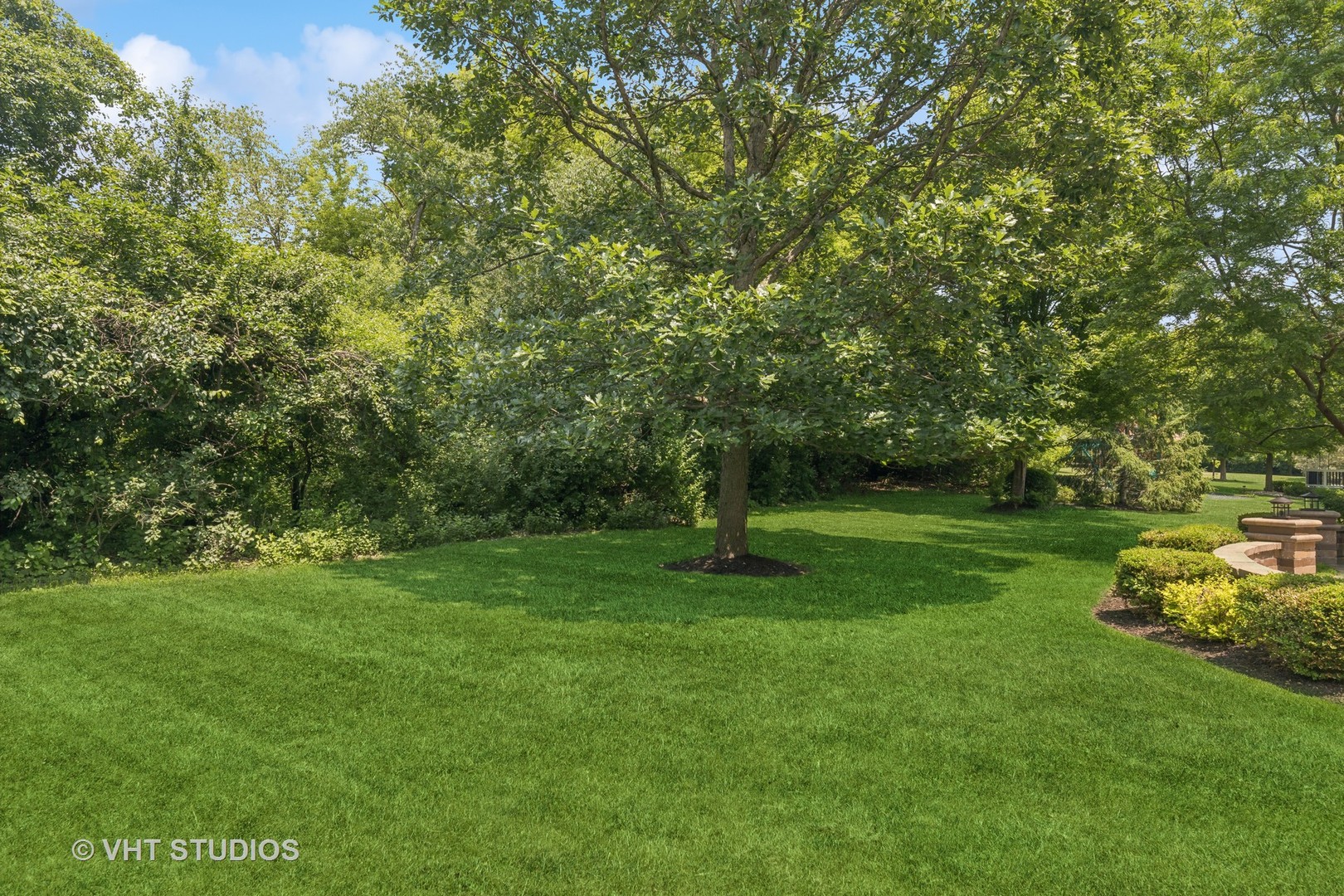 Shaded backyard lawn with mature trees and a brick fire pit at the edge.