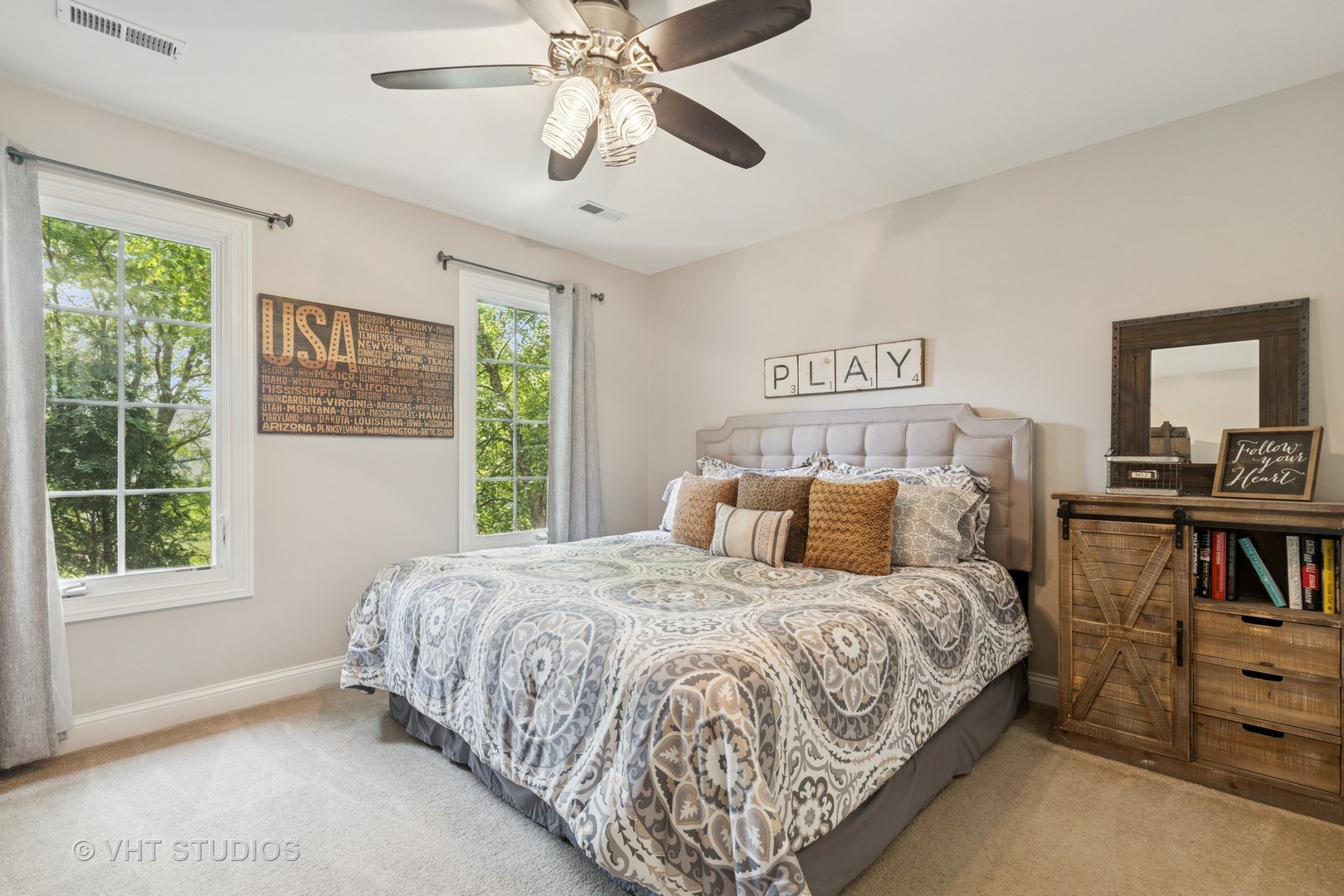 Furnished bedroom with a patterned bedspread, two sunny windows, and a ceiling fan.
