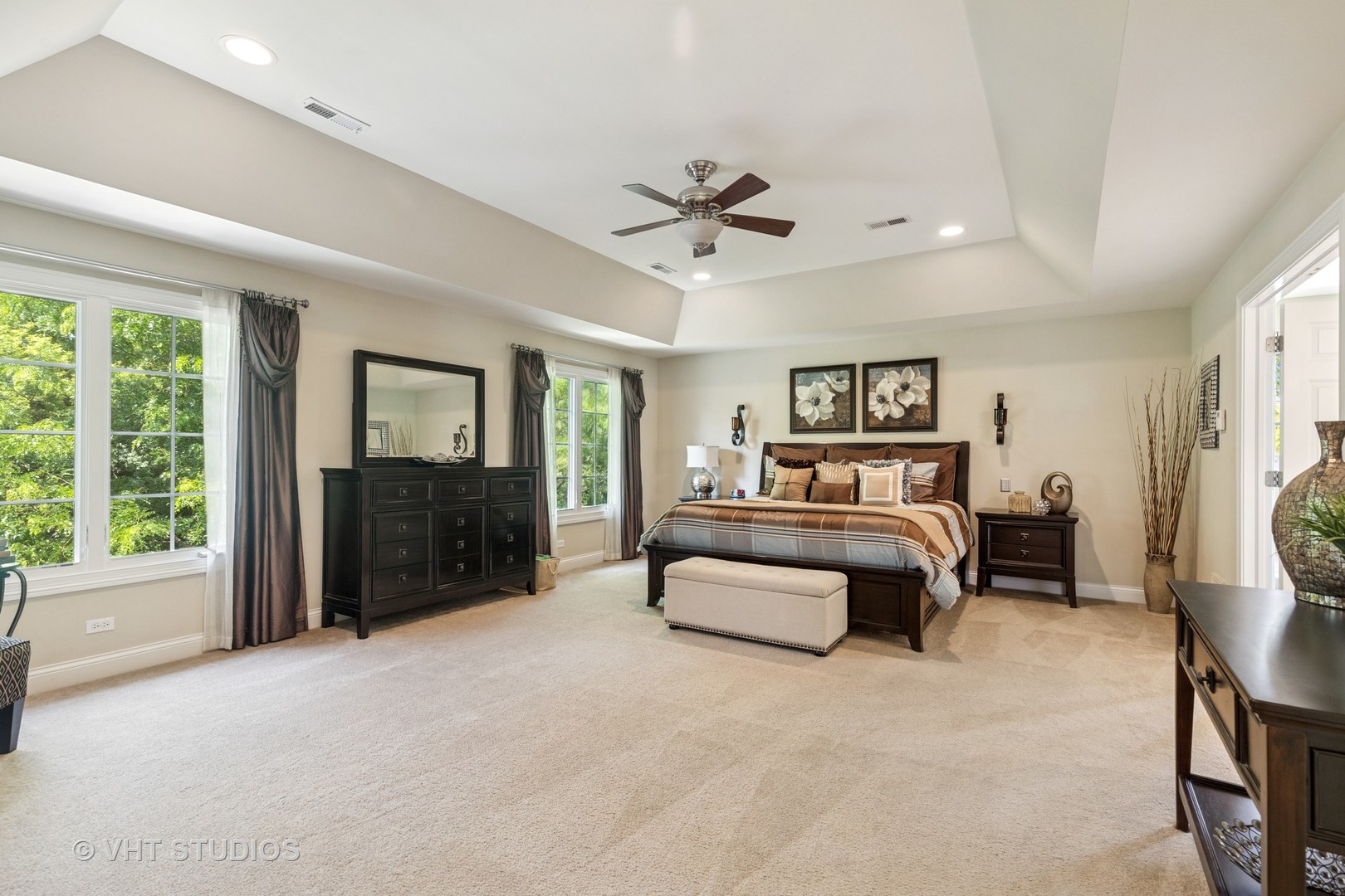 Spacious primary bedroom with a tray ceiling, king-size bed, dark wood furniture, and large windows.