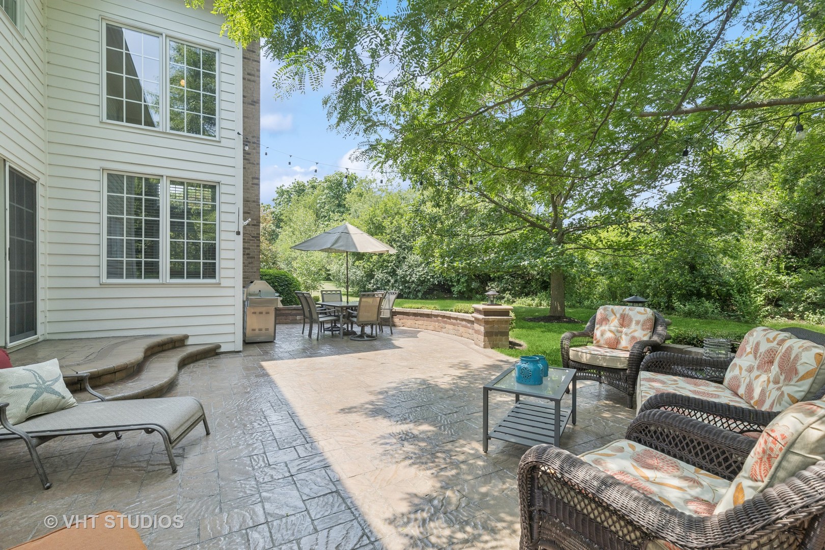 Stone patio with wicker seating beneath a large shade tree, dining table, umbrella, and grill.
