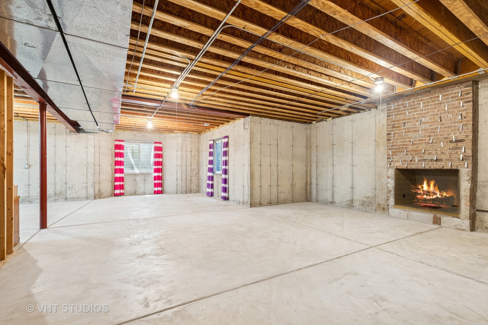 Unfinished basement with exposed ceiling joists, concrete floors, and a brick fireplace with a fire.