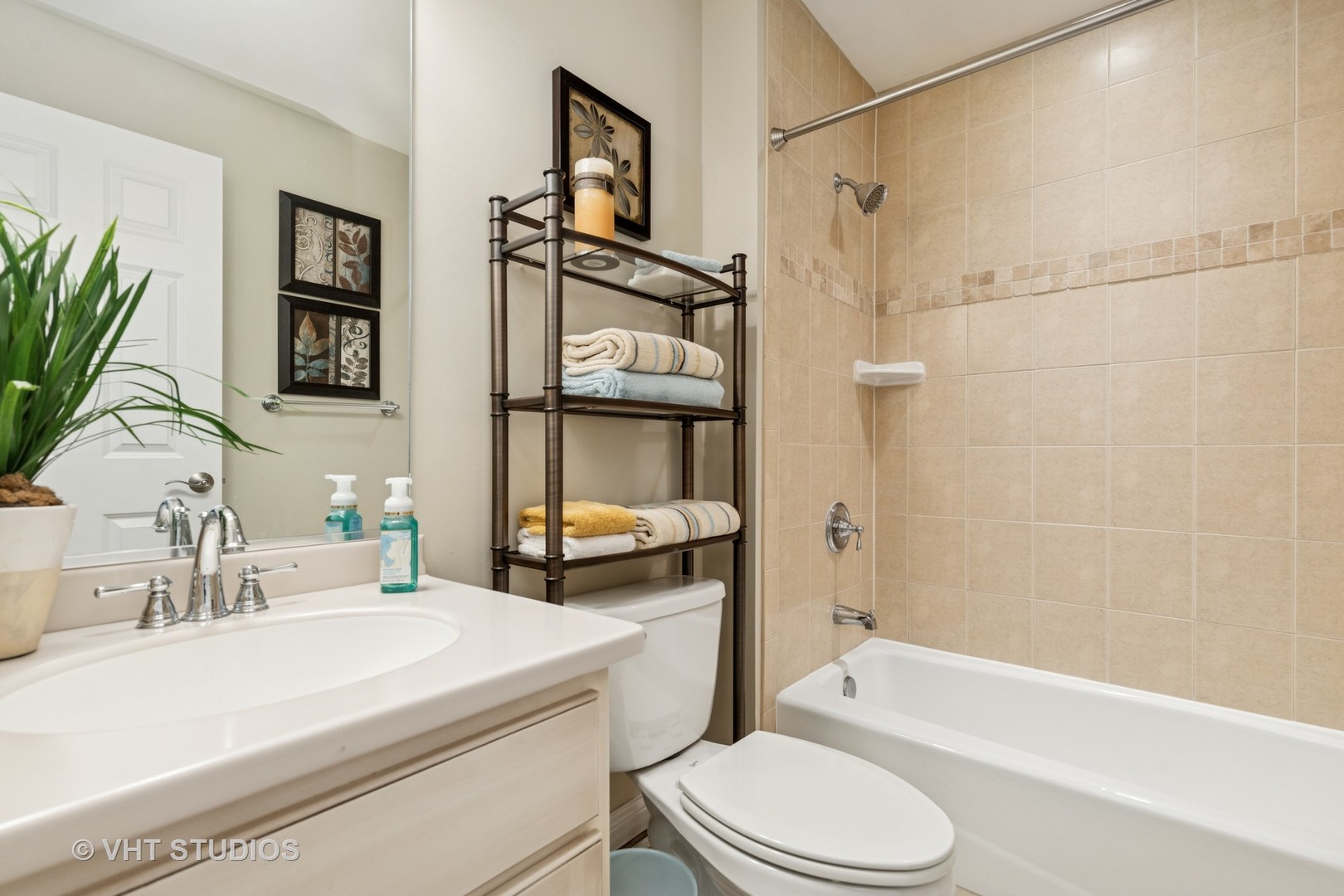 Bathroom with beige tiled tub and shower, white vanity, and folded towels on a metal shelf.