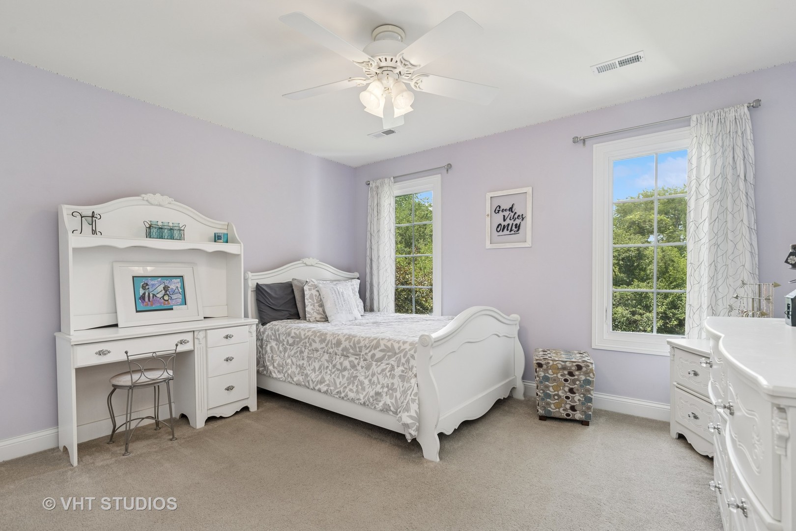 Furnished bedroom with lavender walls, a white bed and desk, and two large curtained windows.