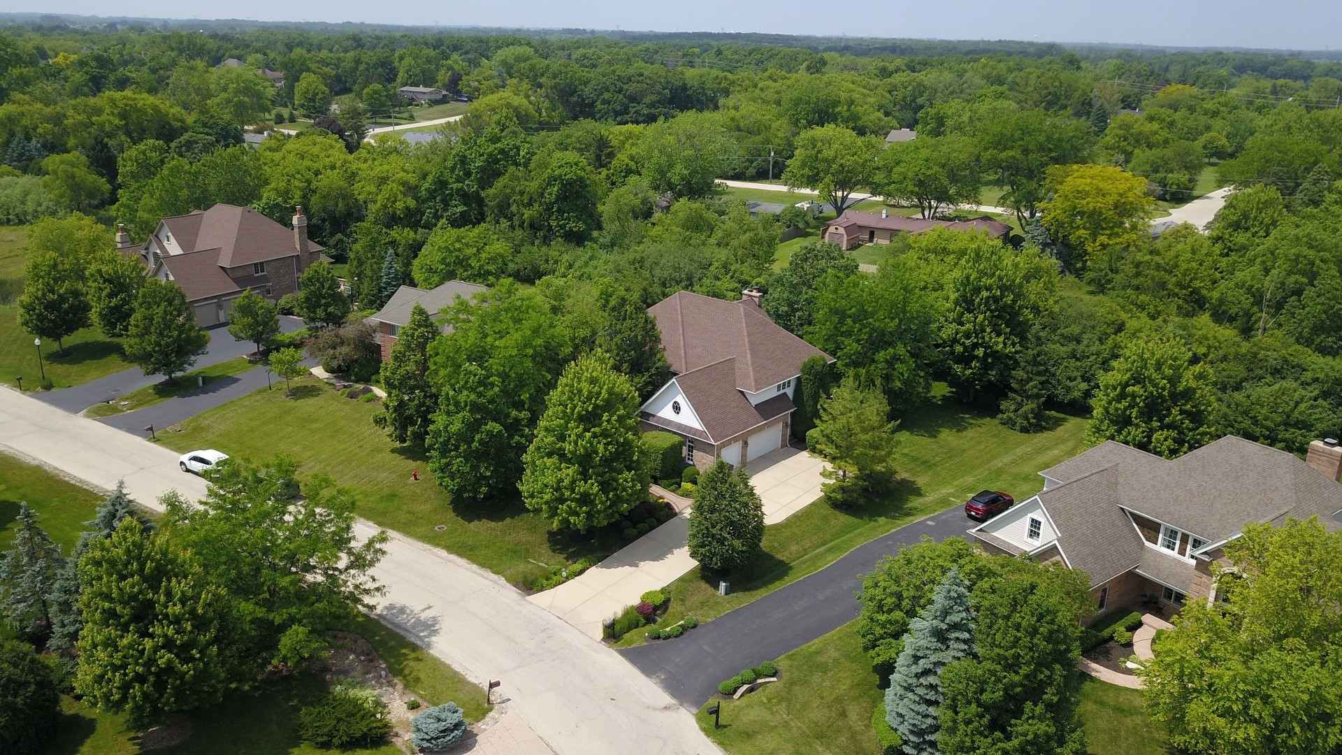 Aerial view of a brick home with a brown roof, attached garage, and tree-lined yard.