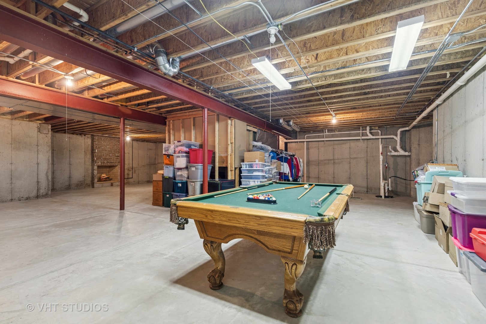 Unfinished basement with a pool table, exposed ceiling joists, storage bins, and a small fireplace.