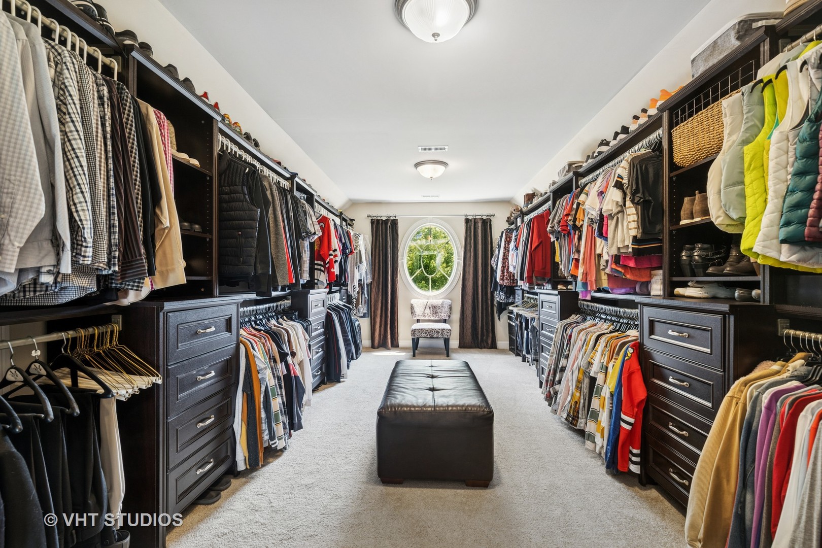Walk-in closet with dark built-in storage, rows of clothing, and a leather bench facing an oval window.